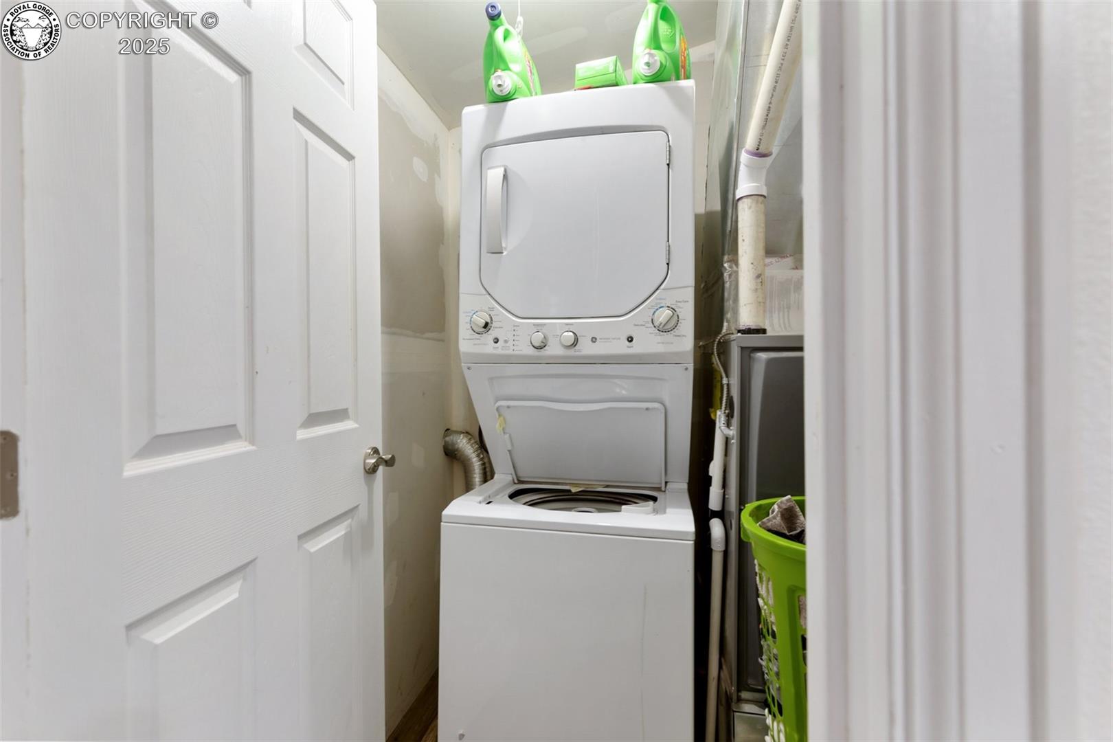 Laundry closet with stacked washer and dryer, open washer lid, and cleaning supplies.