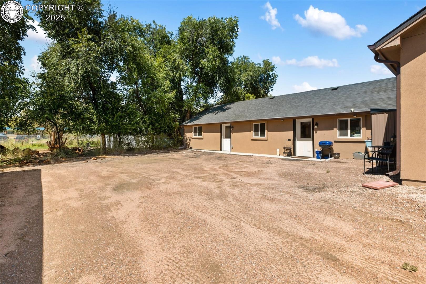 Single-story tan home with a broad gravel yard, mature trees, outdoor chairs, and a barbecue grill.