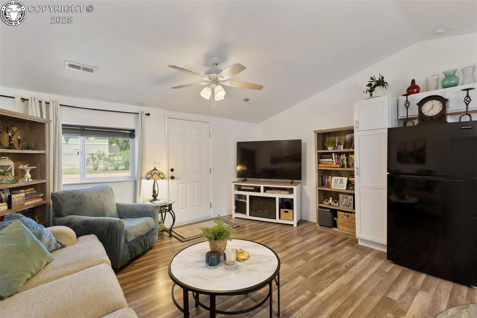 Furnished living room with wood-look floors, a round coffee table, ceiling fan, and large black refrigerator.