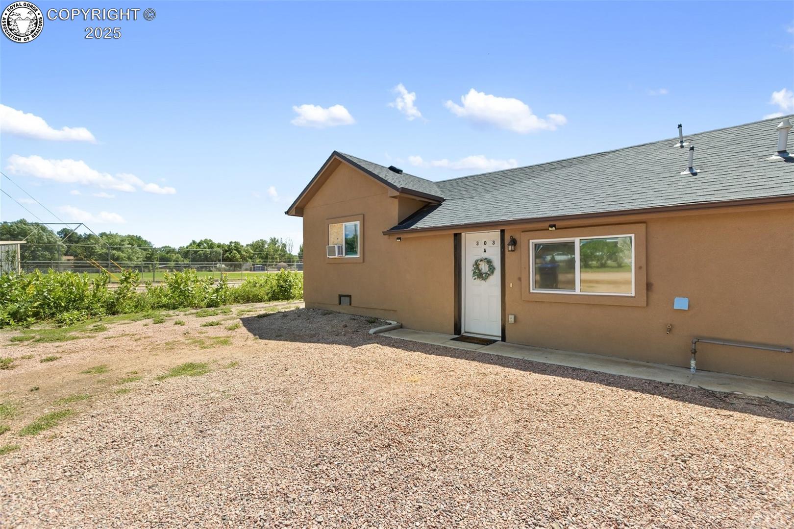 Tan stucco home with a gravel yard, white entry door, and fenced green area beside it.