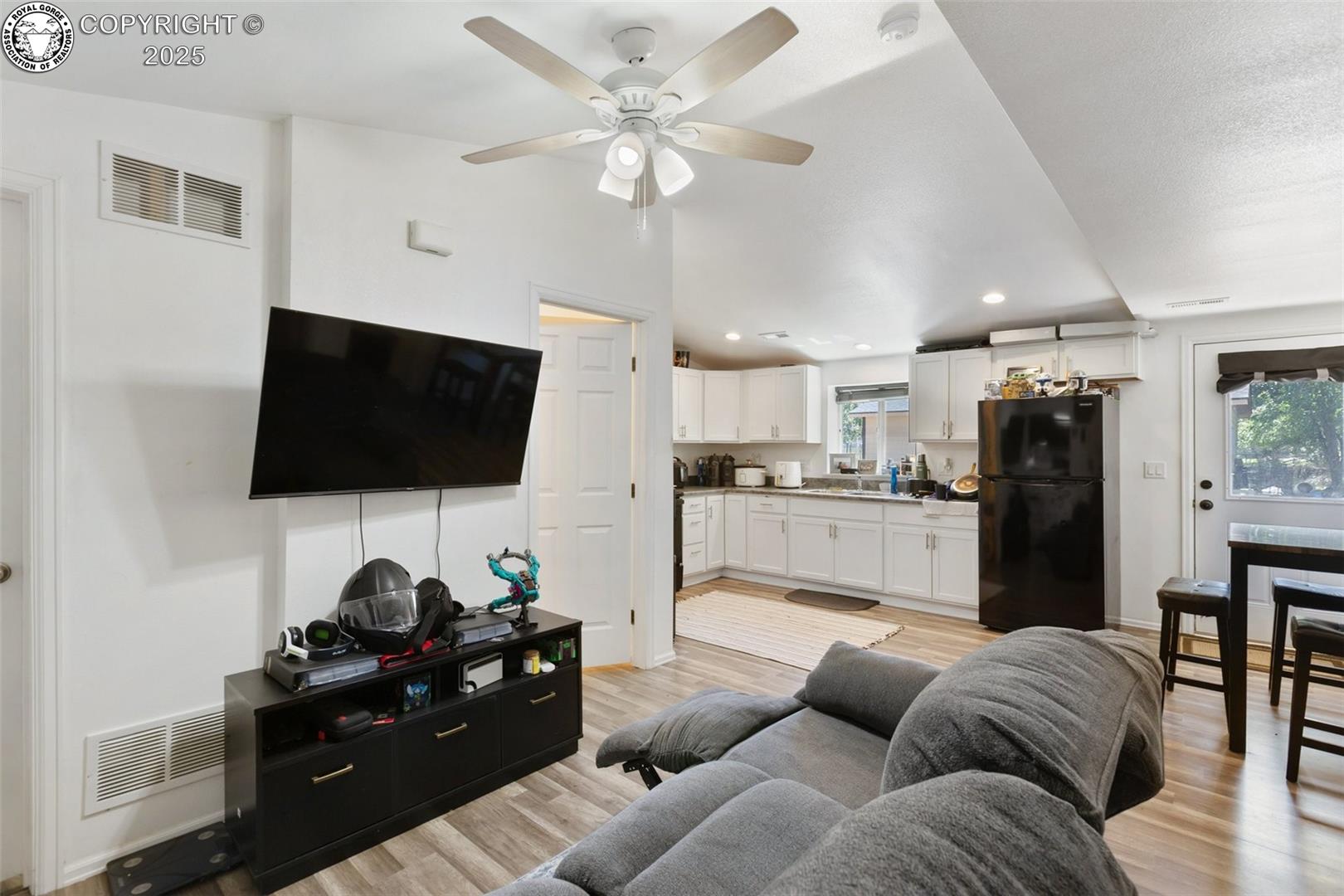 Living room with a gray sectional and wall-mounted television opening onto a bright kitchen.