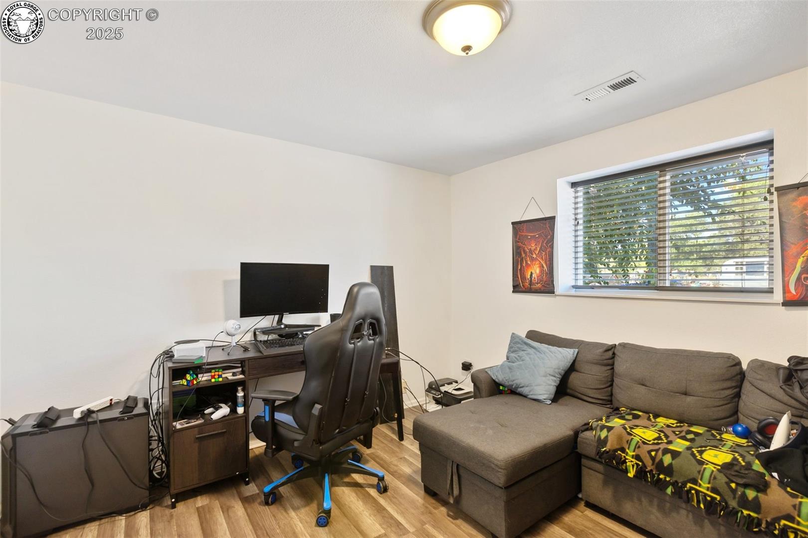 Furnished room with a computer desk and gaming chair beside a gray sectional sofa and wide window.