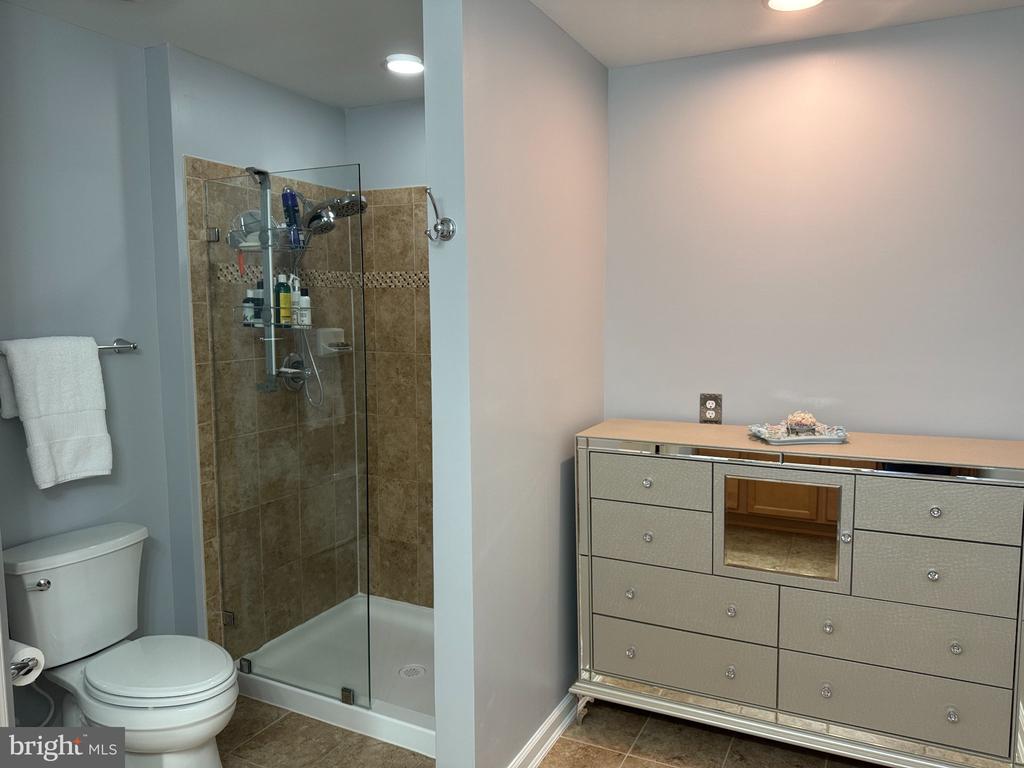 Bathroom with a glass-enclosed tan-tile shower, toilet, and wide silver dresser.