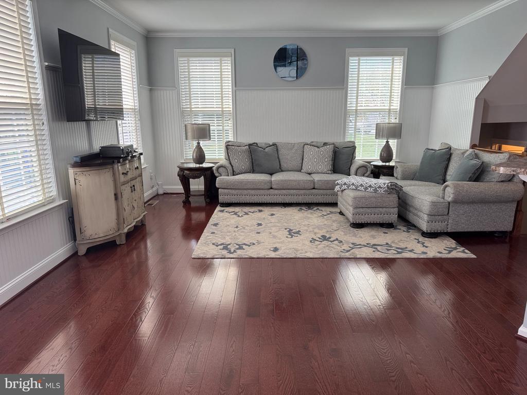 Living room with gray sofas, a patterned rug, dark hardwood floors, and tall windows with blinds.