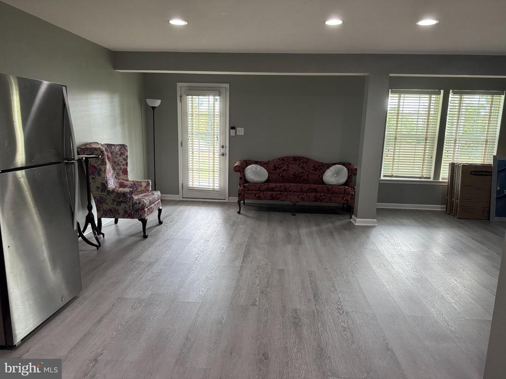 Living area with gray flooring, floral seating, a stainless-steel refrigerator, and bright windows with blinds.