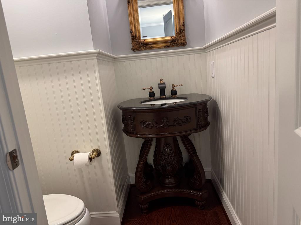 Powder room with a dark ornate pedestal vanity, brass fixtures, white beadboard walls, and a framed mirror.