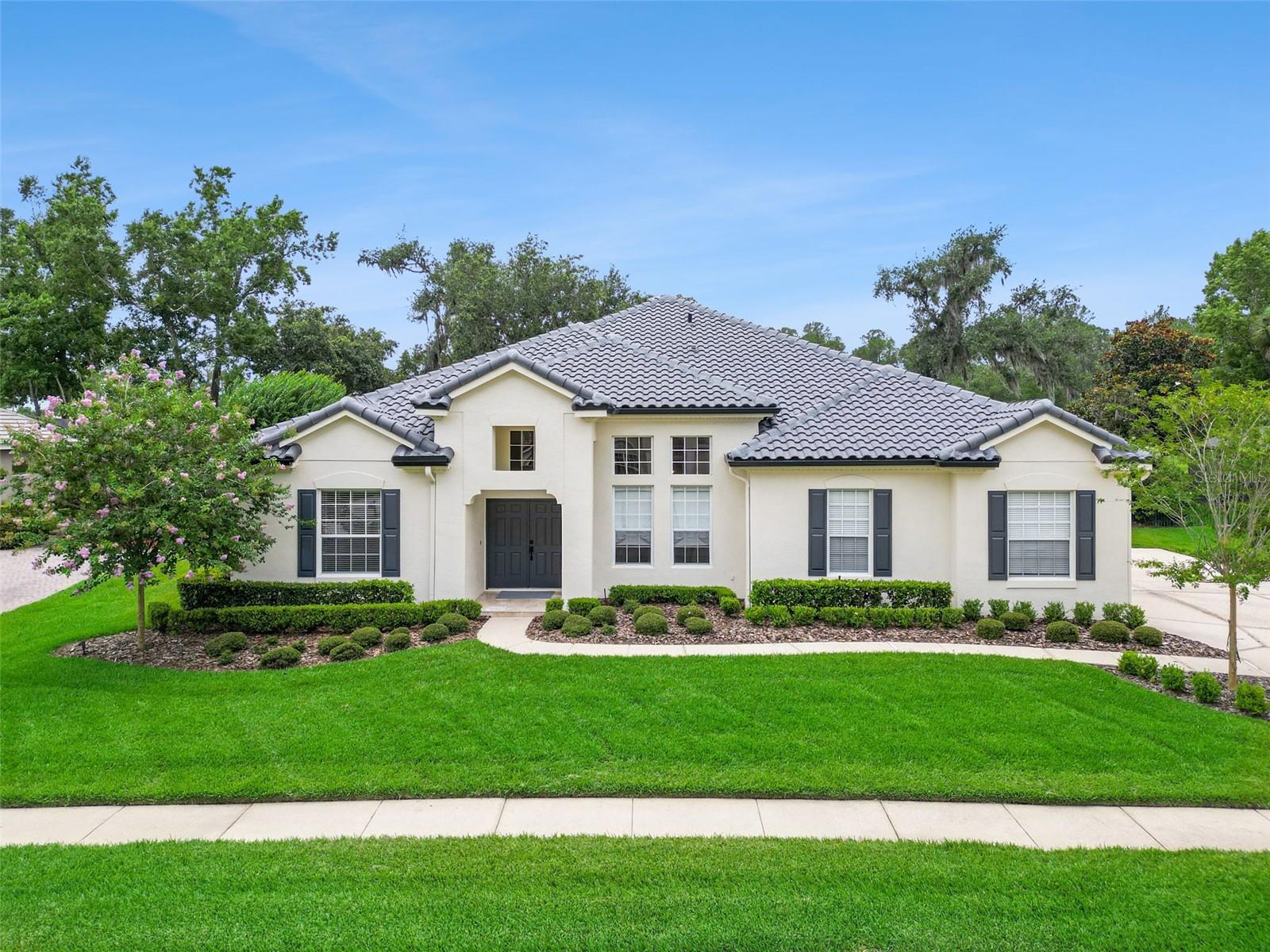 Single-story stucco home with a gray tile roof, dark shutters, and neatly landscaped front lawn.
