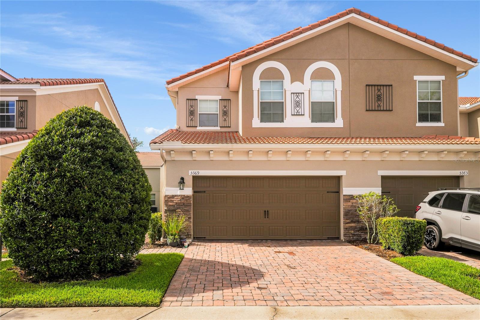 Two-story tan stucco home with a tile roof, arched windows, and a brick-paver driveway.