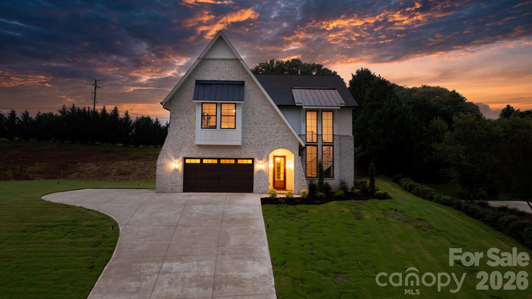 Two-story brick home with a steep gable and glowing windows at dusk, viewed from its curved driveway.