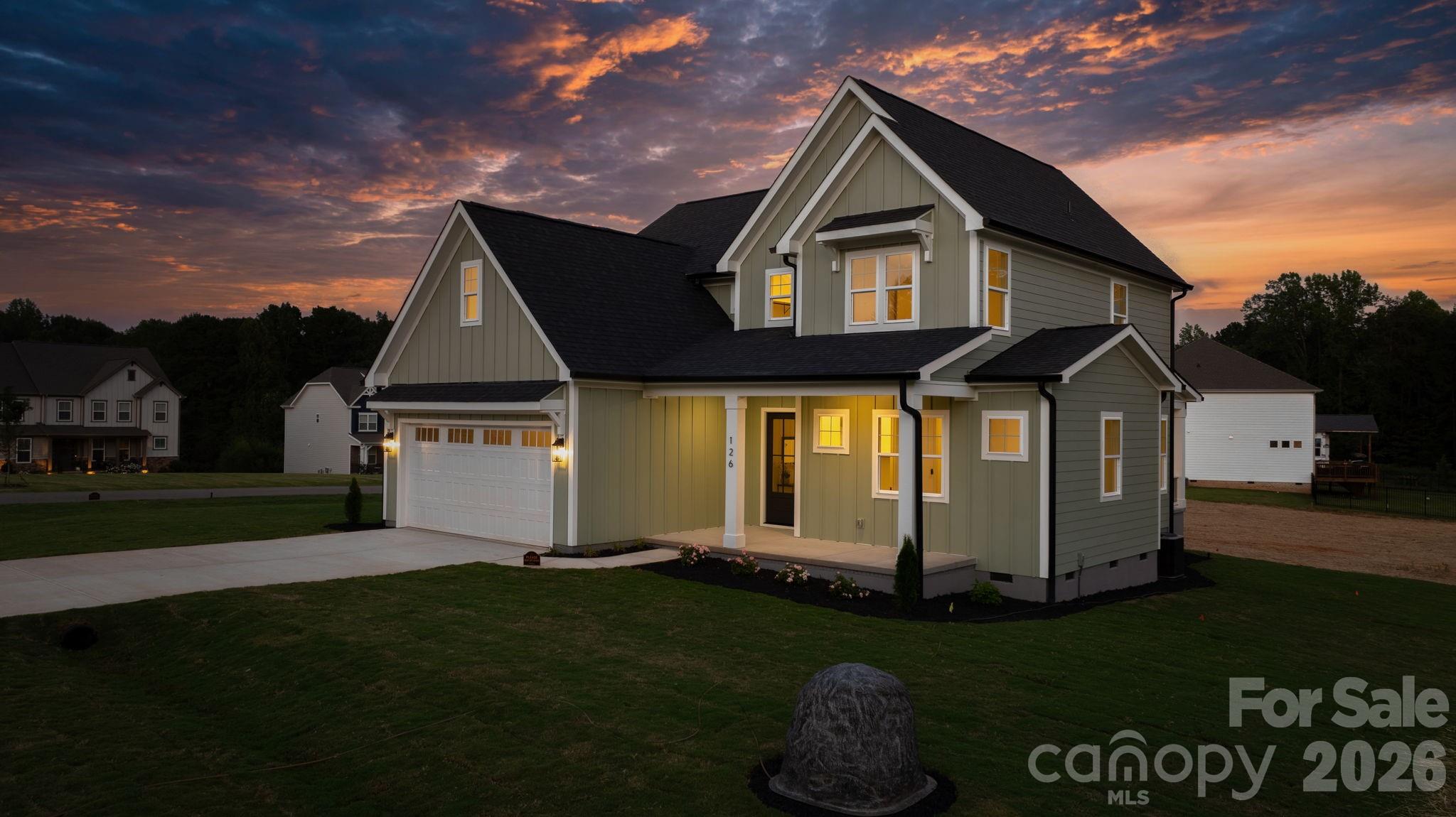 Two-story gray-green house with a double garage and warmly lit windows beneath a colorful dusk sky.