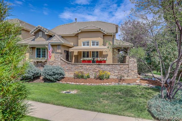 Two-story stucco home with a tile roof, stone garden wall, American flag, and landscaped lawn.