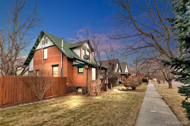 Red-brick Denver home with a steep green roof, wooden fence, and sidewalk in front.