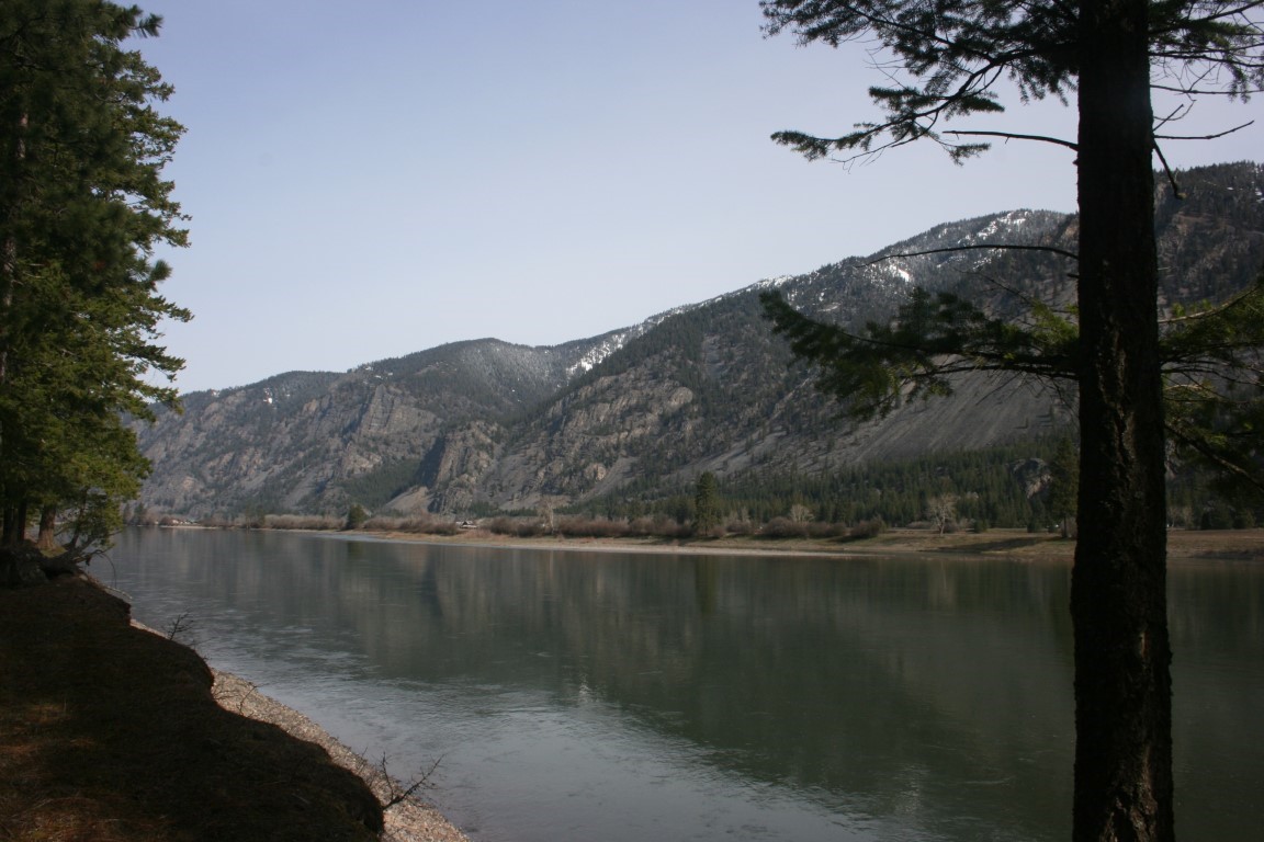 Riverfront shoreline framed by evergreens, with calm water and snow-patched mountains beyond.