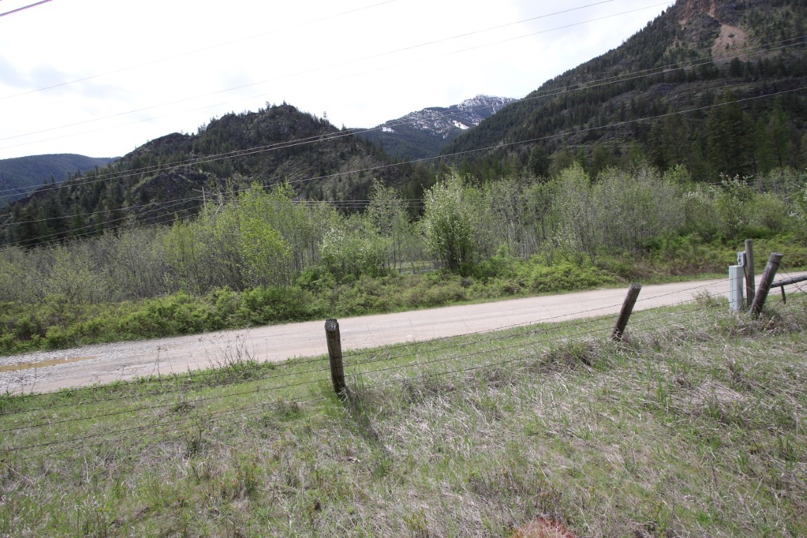 Rural land beside a gravel road and wire fence, backed by forested mountains with patches of snow.