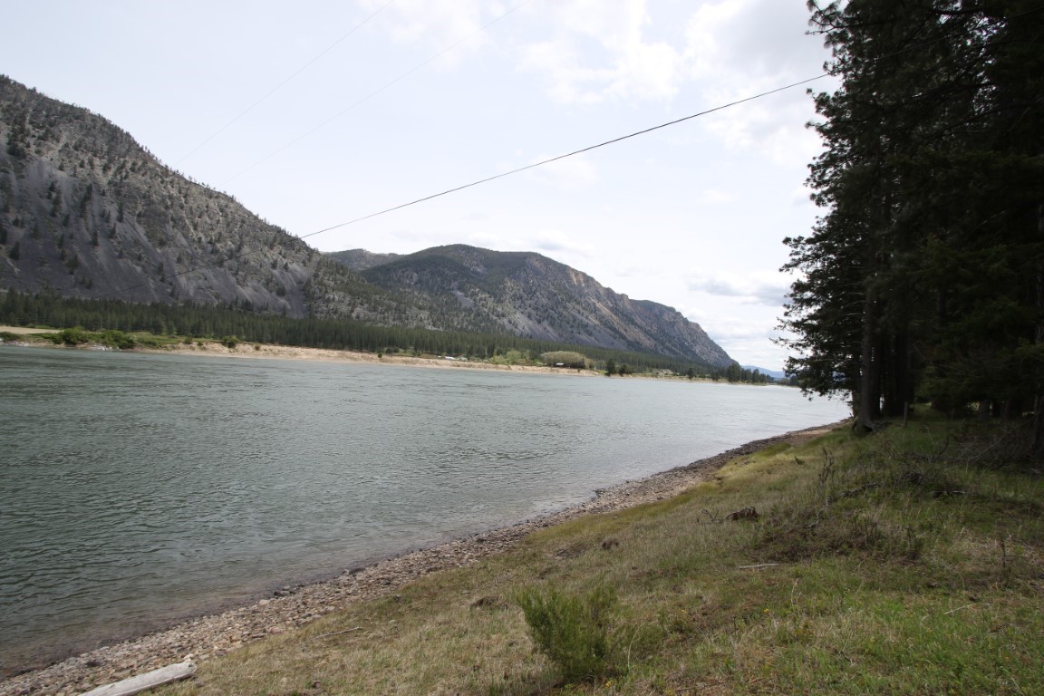 Riverfront land with a pebbled shore, pine trees, and rugged mountains across the water.