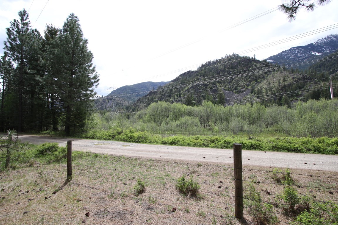 Vacant land beside a gravel road, with a wire fence, pine trees, and wooded mountains beyond.
