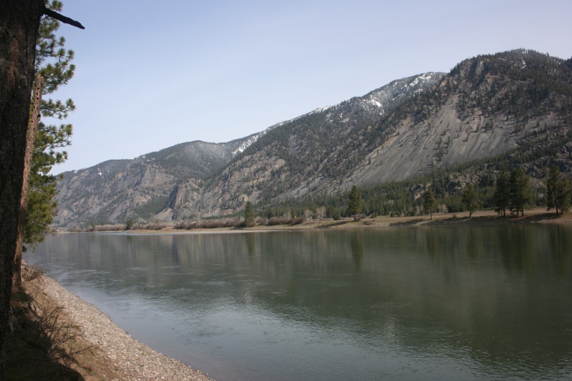 Riverfront shoreline with calm water and steep, partly snow-covered mountains beyond.