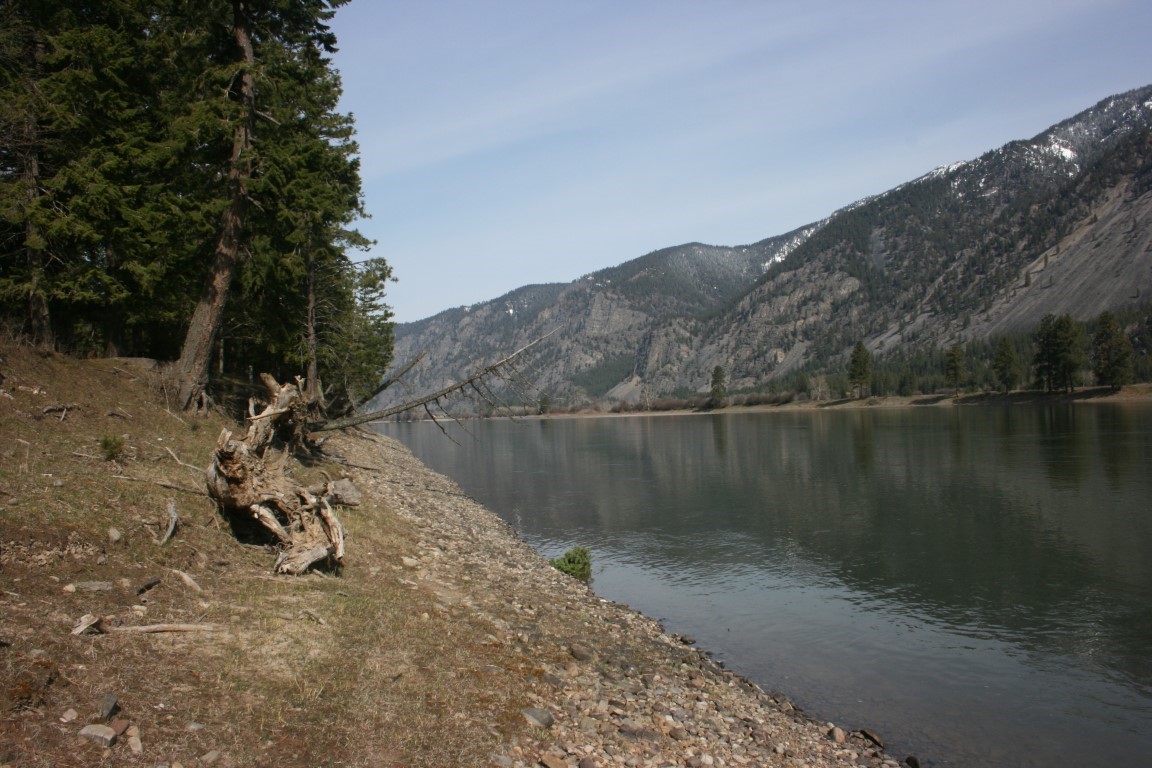 Riverfront land with a rocky, tree-lined shore, calm water, and snow-patched mountains beyond.