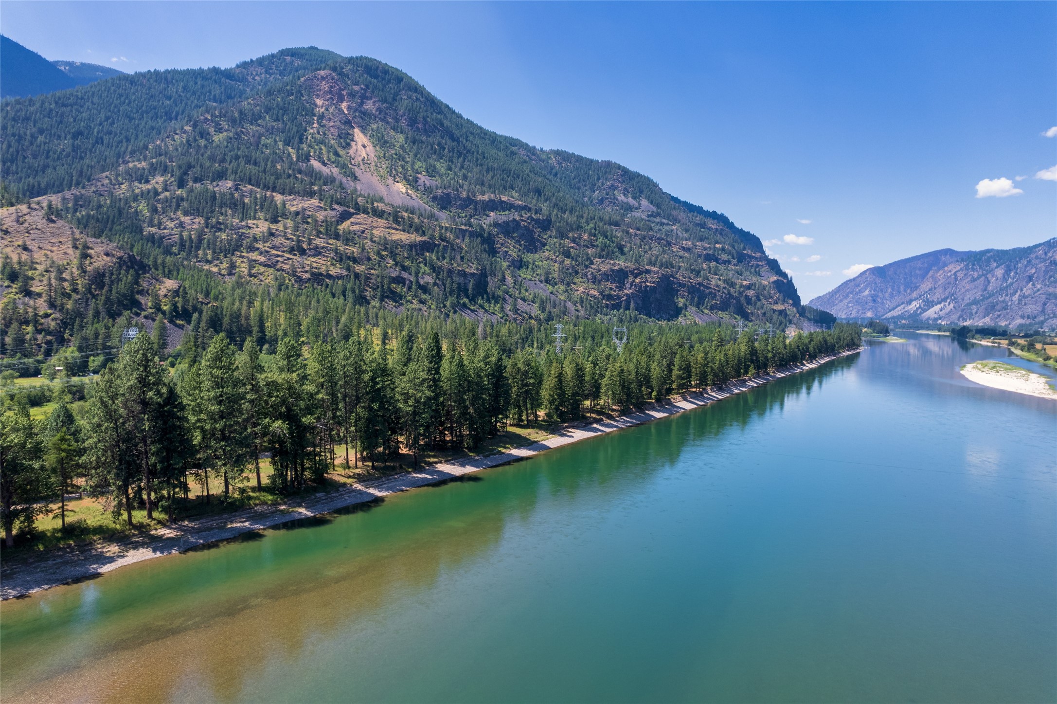 Riverfront land with a tree-lined bank, green-blue water, and forested mountains.