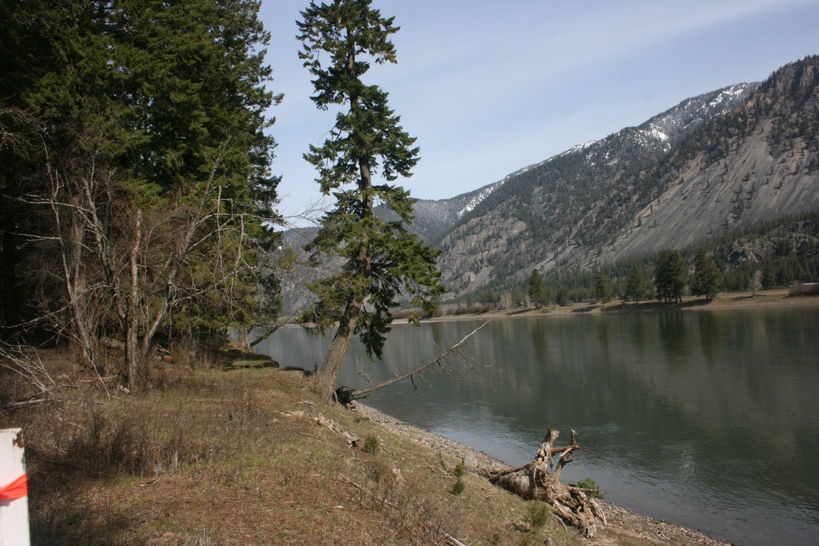 Riverfront property with a wooded shoreline, calm water, and a steep mountain across the river.