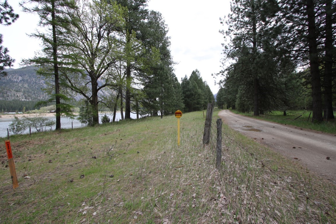 Vacant riverside land beside a tree-lined dirt road, with distant mountains and boundary markers.