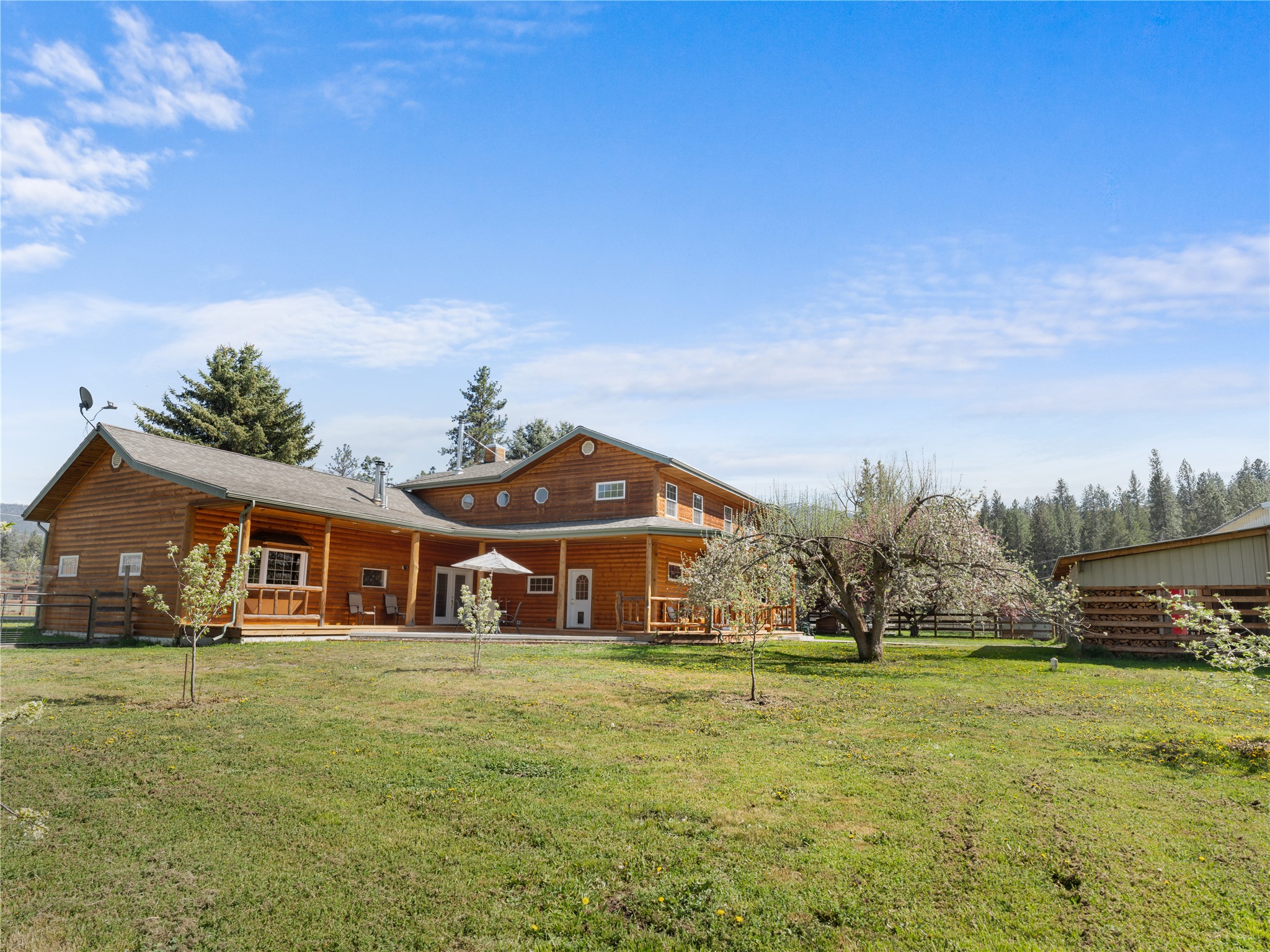 Wooden house with a long covered porch, flowering trees, and a woodshed in a grassy yard.
