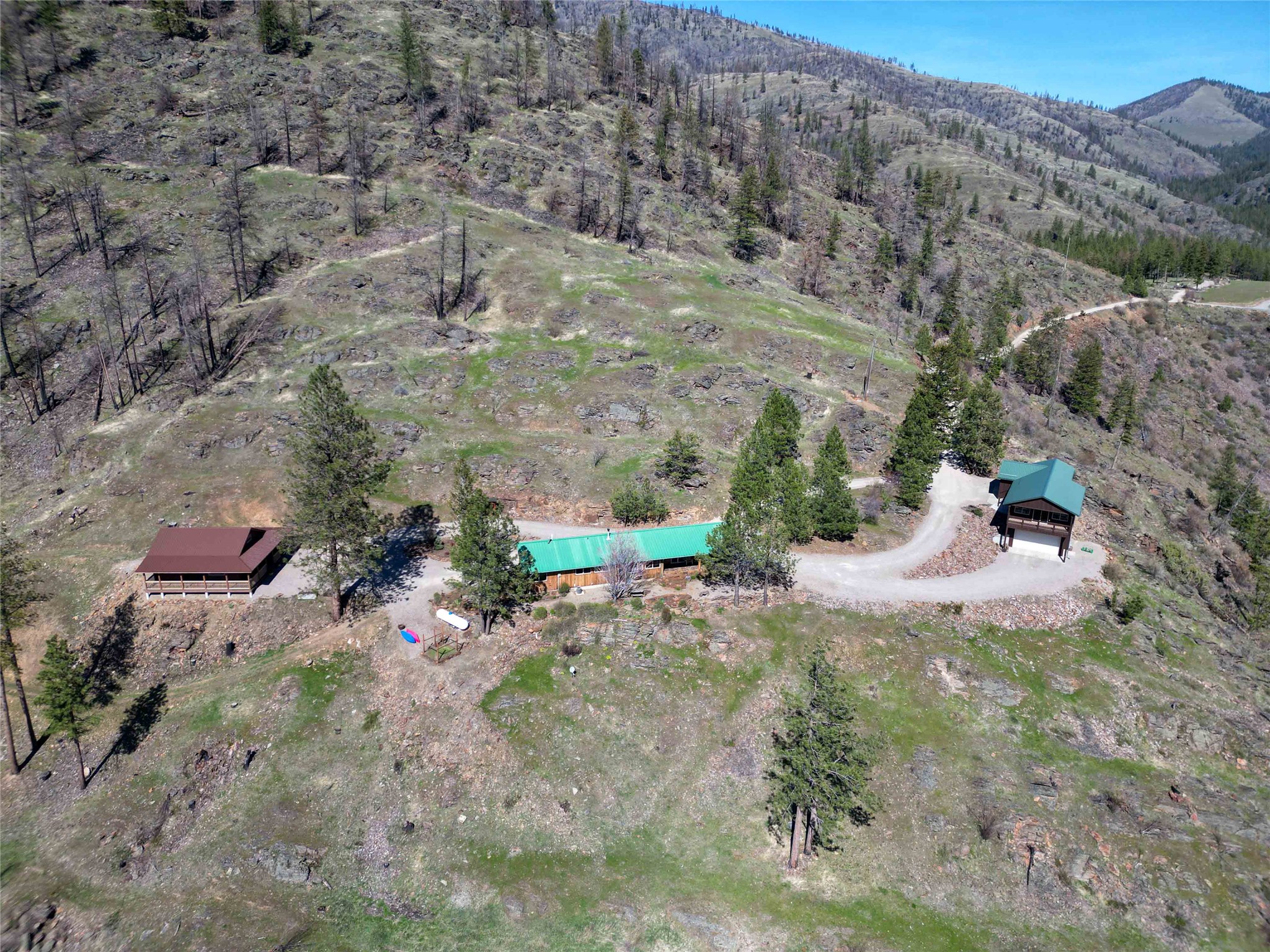 Aerial view of a mountainside property with three buildings and a curved gravel driveway.