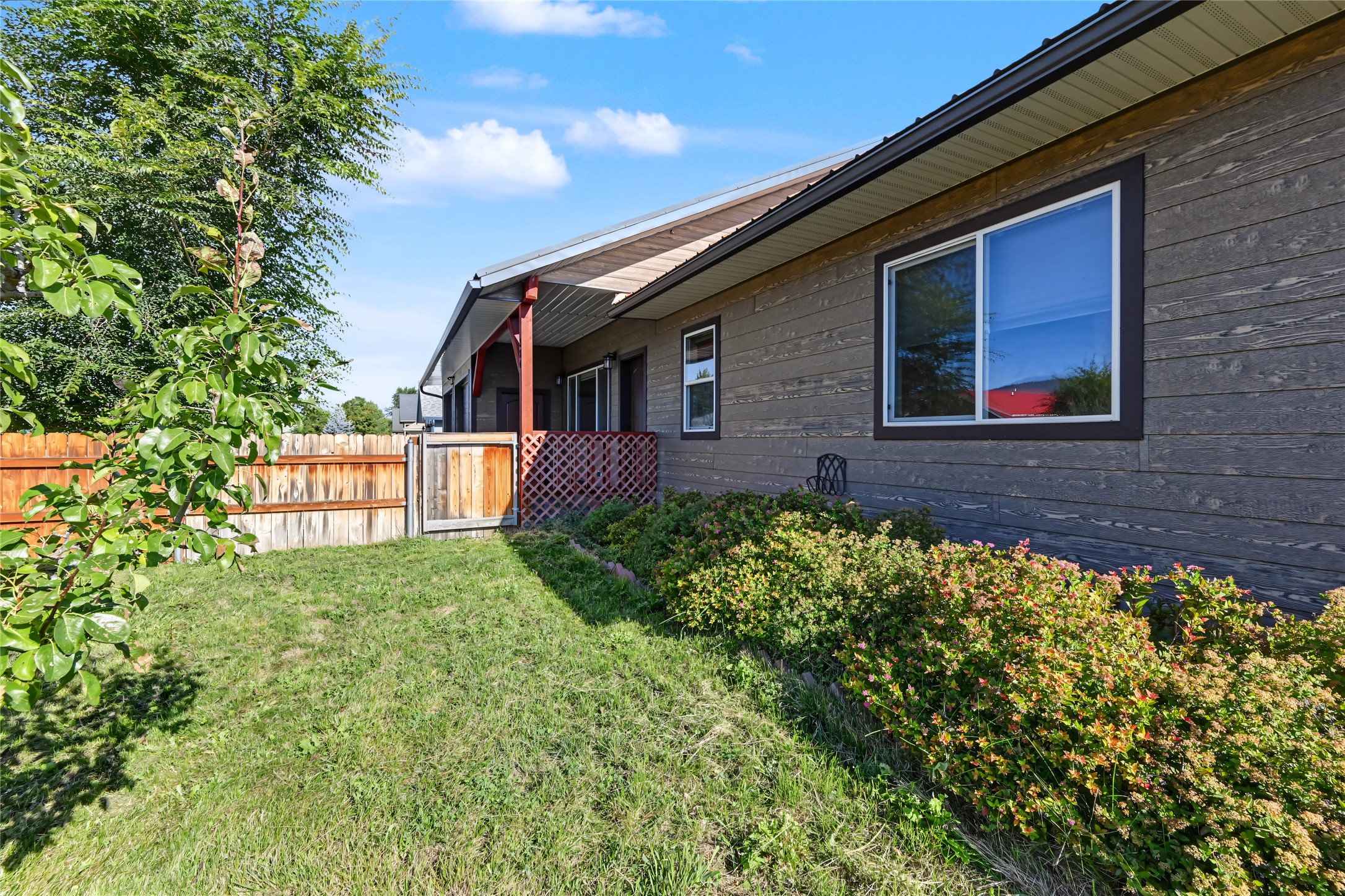 House side with dark wood siding, large windows, flowering shrubs, and a fenced grassy yard.