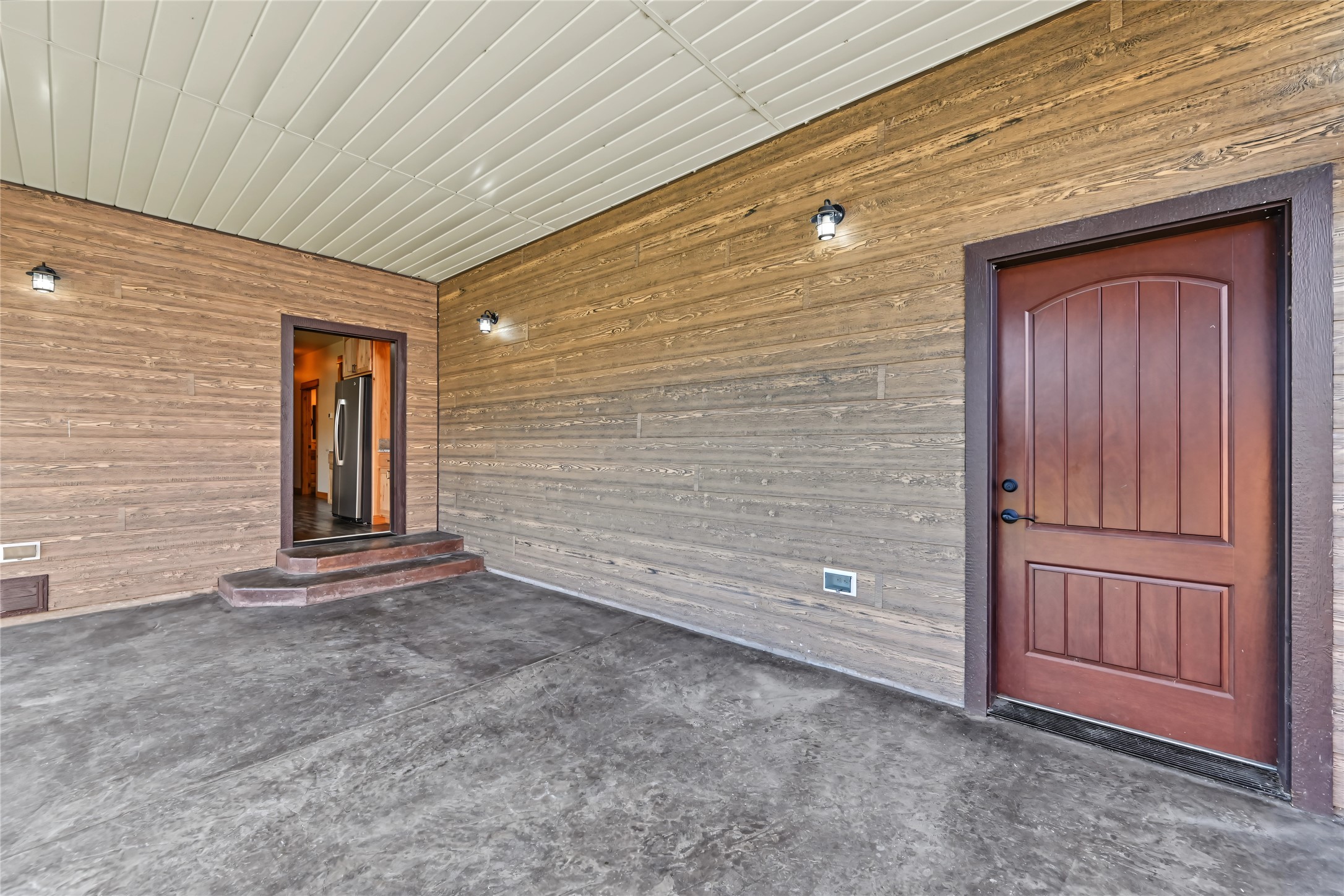 Covered porch with weathered wood siding, concrete floor, and a reddish-brown front door.