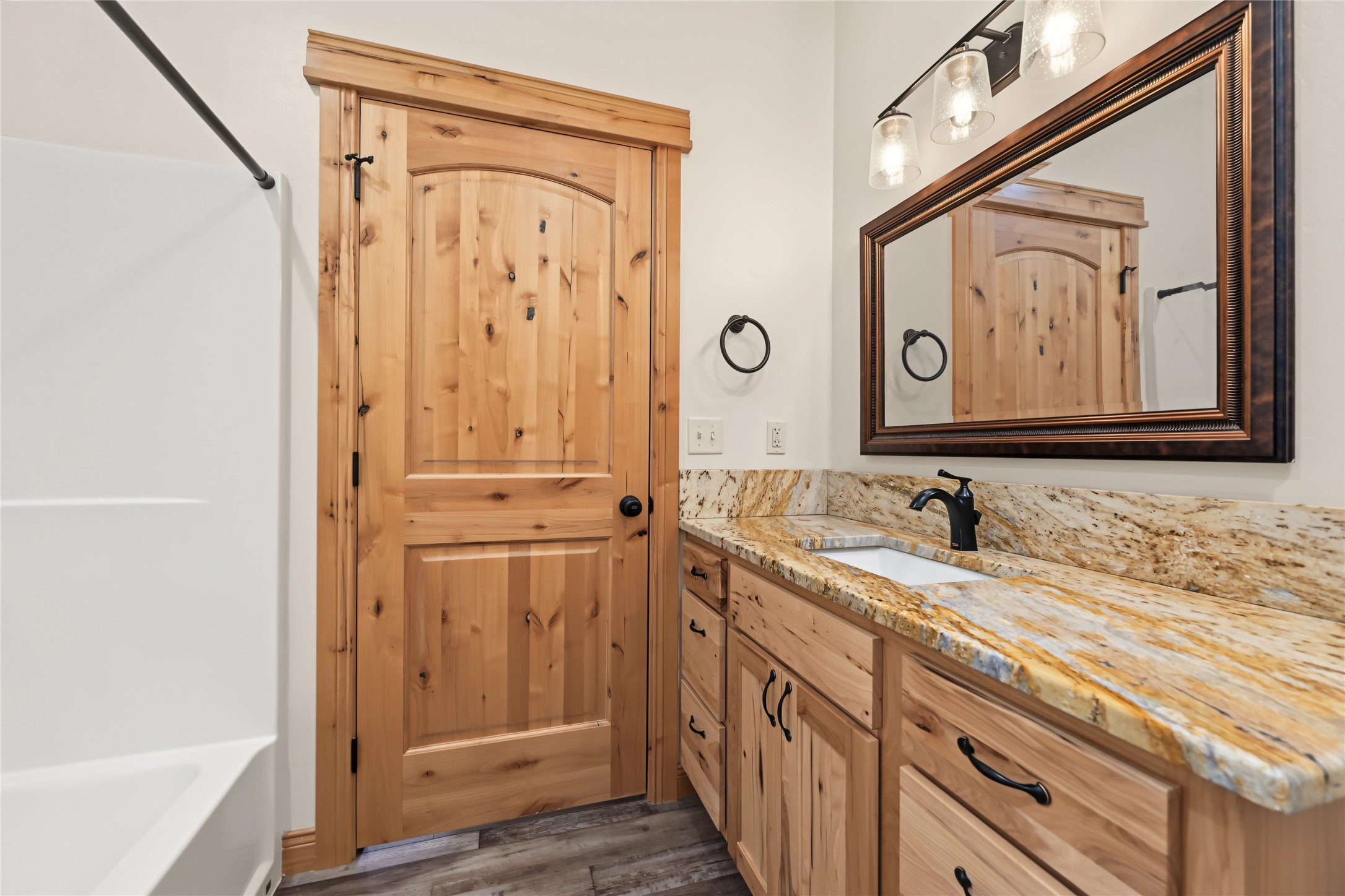 Bathroom vanity with knotty pine cabinetry, stone countertop, dark-framed mirror, and a white bathtub.
