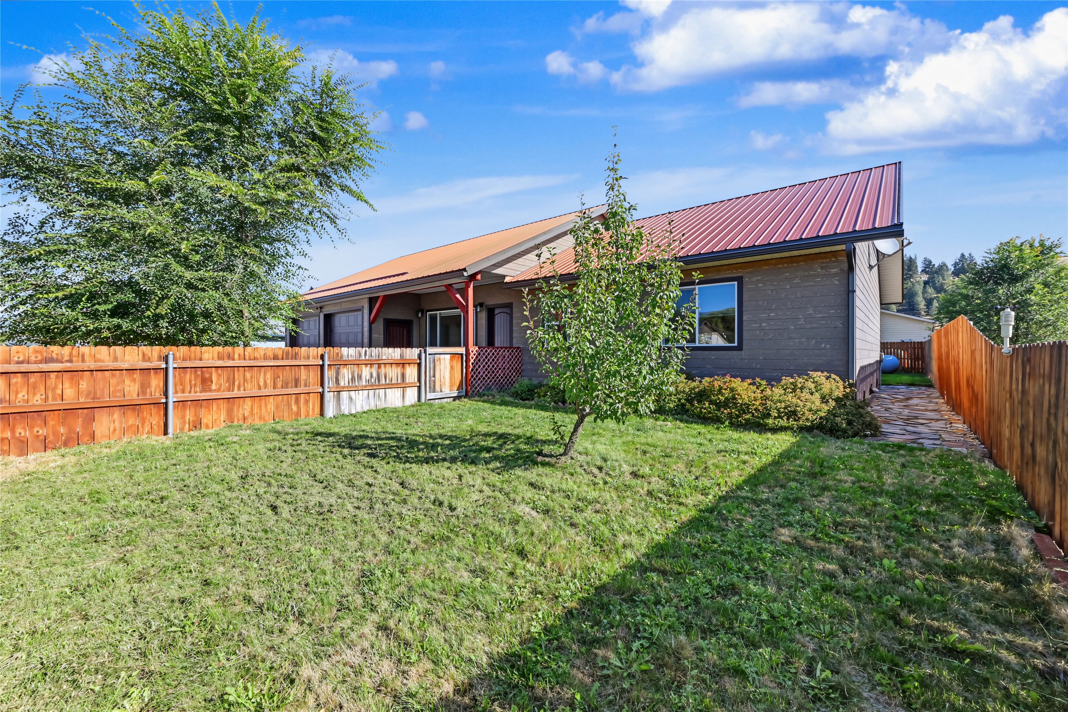 Single-story home with a red metal roof behind a fenced grassy yard and young trees.