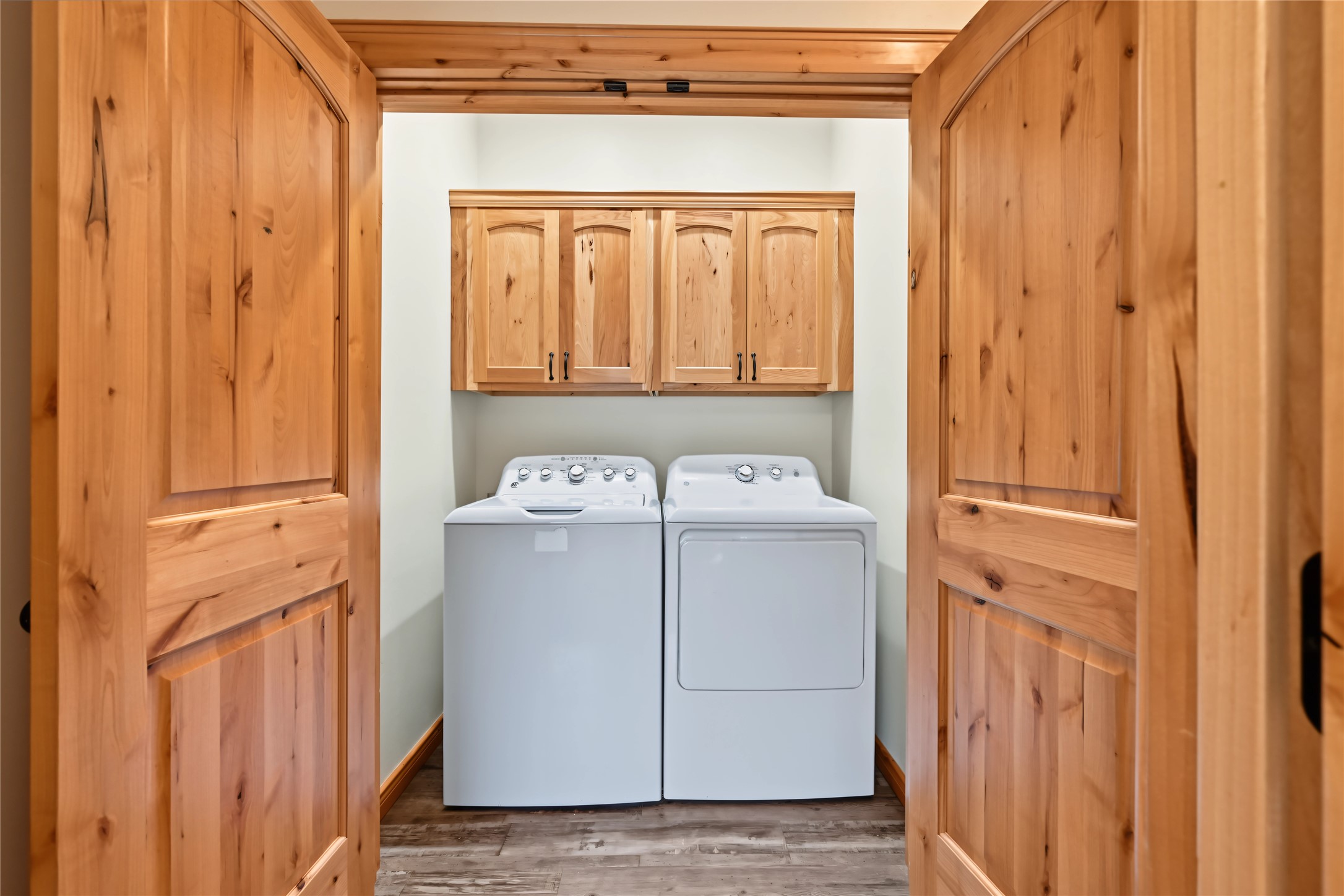 Laundry room with a white washer and dryer beneath natural wood cabinets, framed by matching wooden doors.
