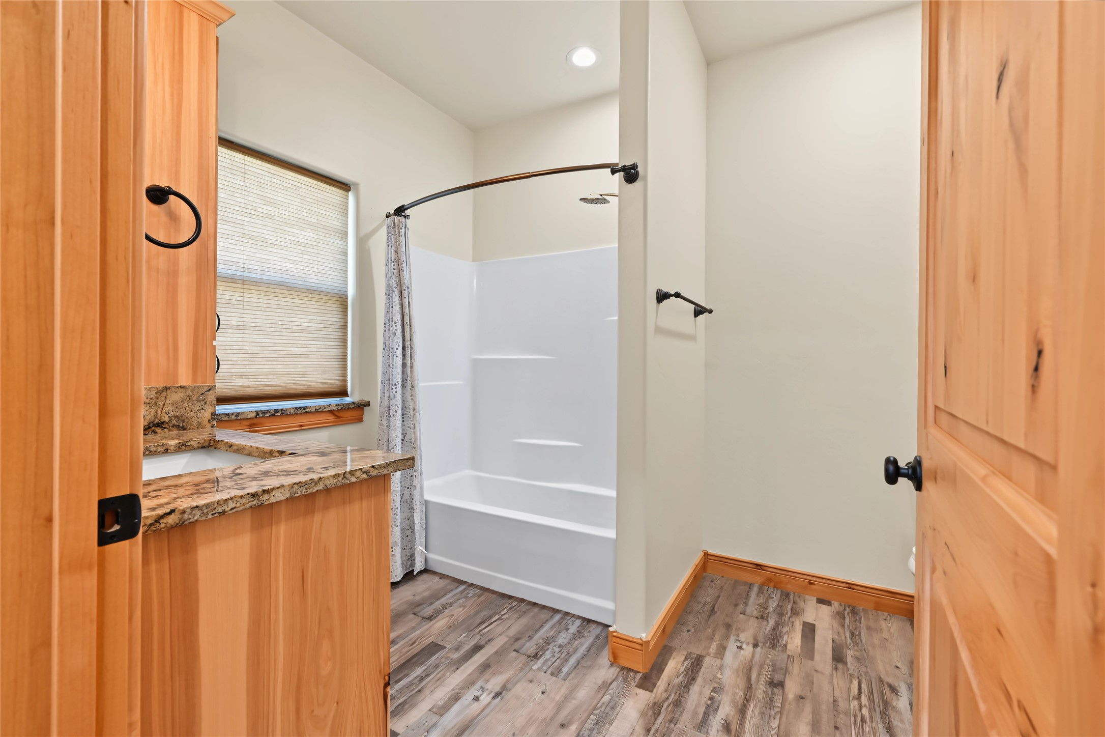 Bathroom with a white tub-shower, granite vanity counter, wood cabinetry, and plank flooring.