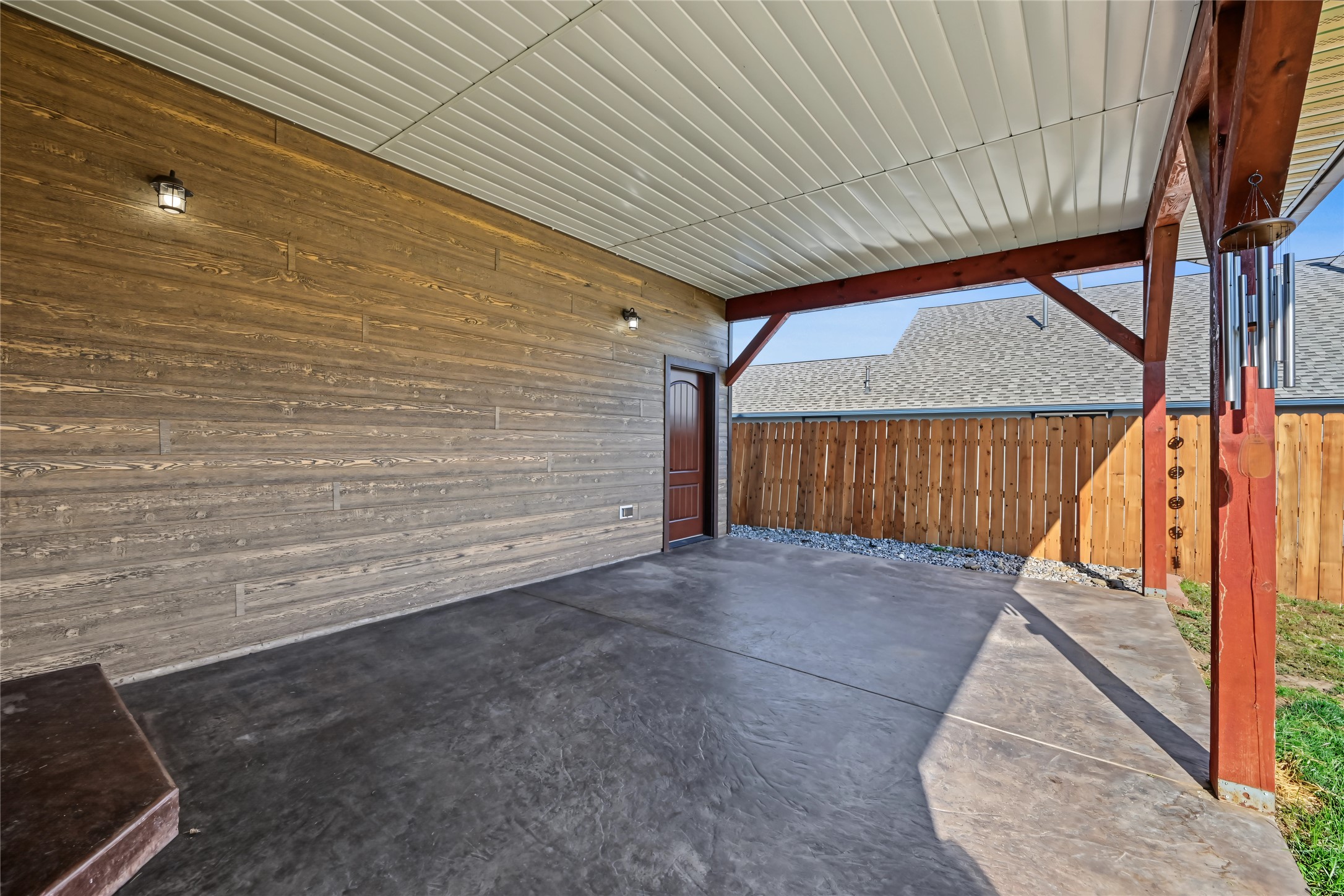 Covered patio with a concrete floor, rustic wood siding, red timber supports, and a wooden privacy fence.