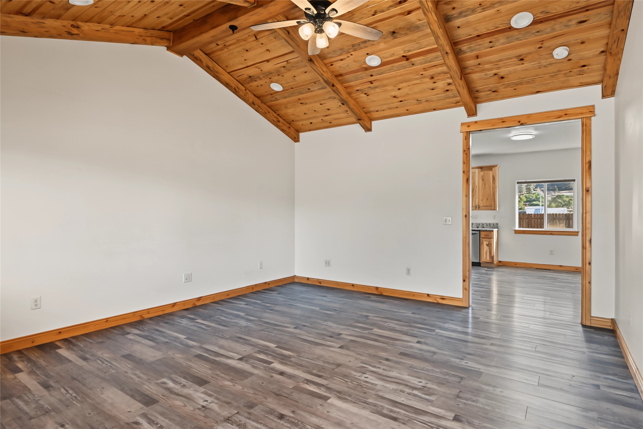 Empty living room with a vaulted wood ceiling and gray plank floors, opening to the kitchen.