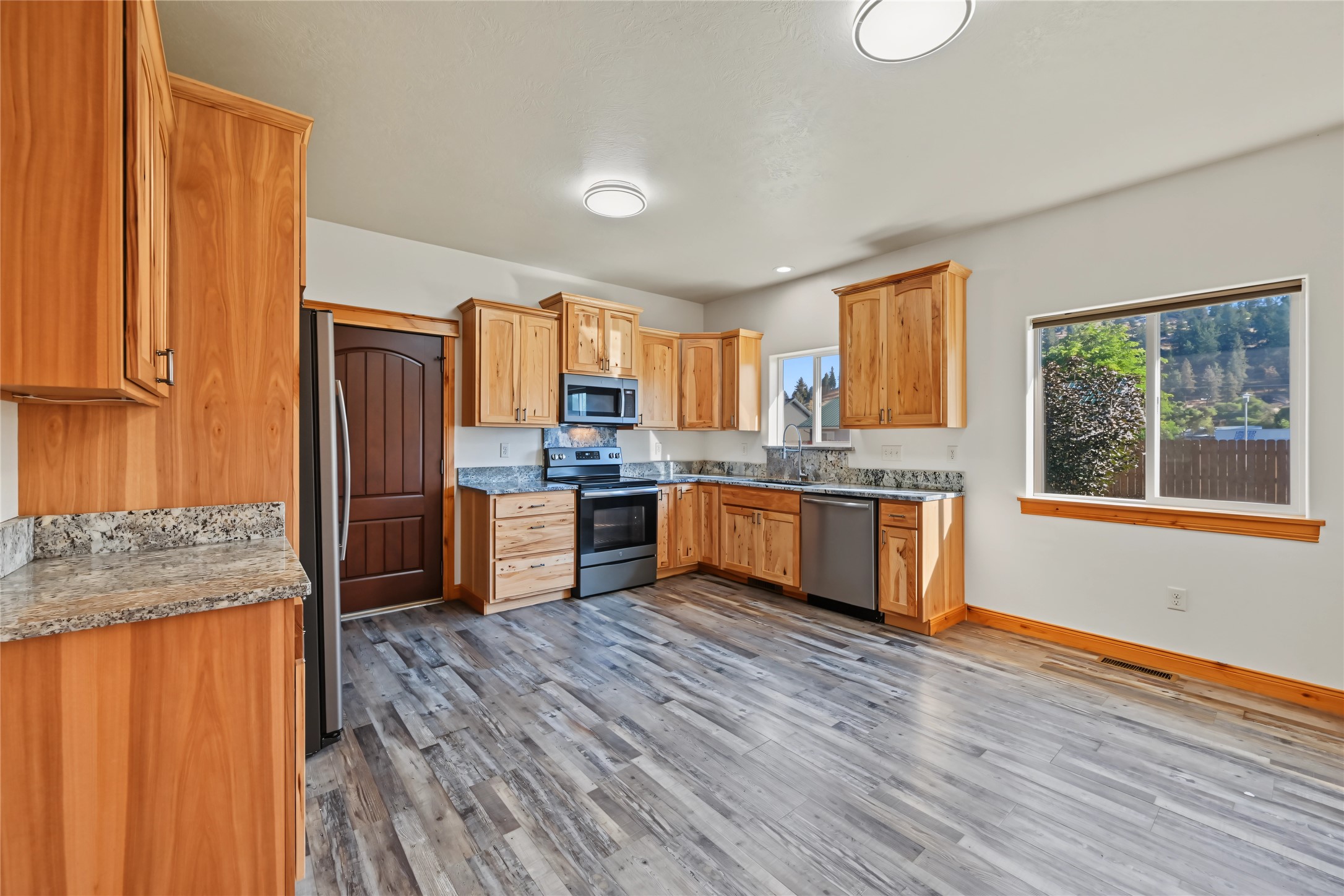 Empty kitchen with honey-colored wood cabinets, granite countertops, stainless steel appliances, and gray flooring.