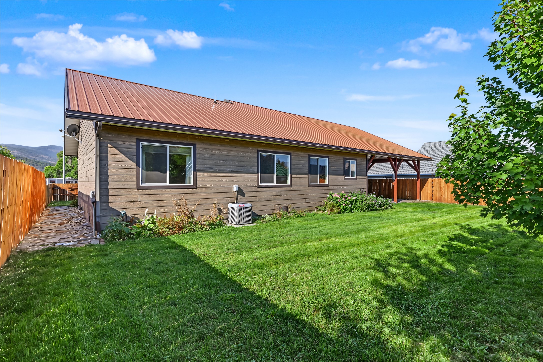Wood-sided home with a rust-colored metal roof, green lawn, fenced walkway, and covered carport.