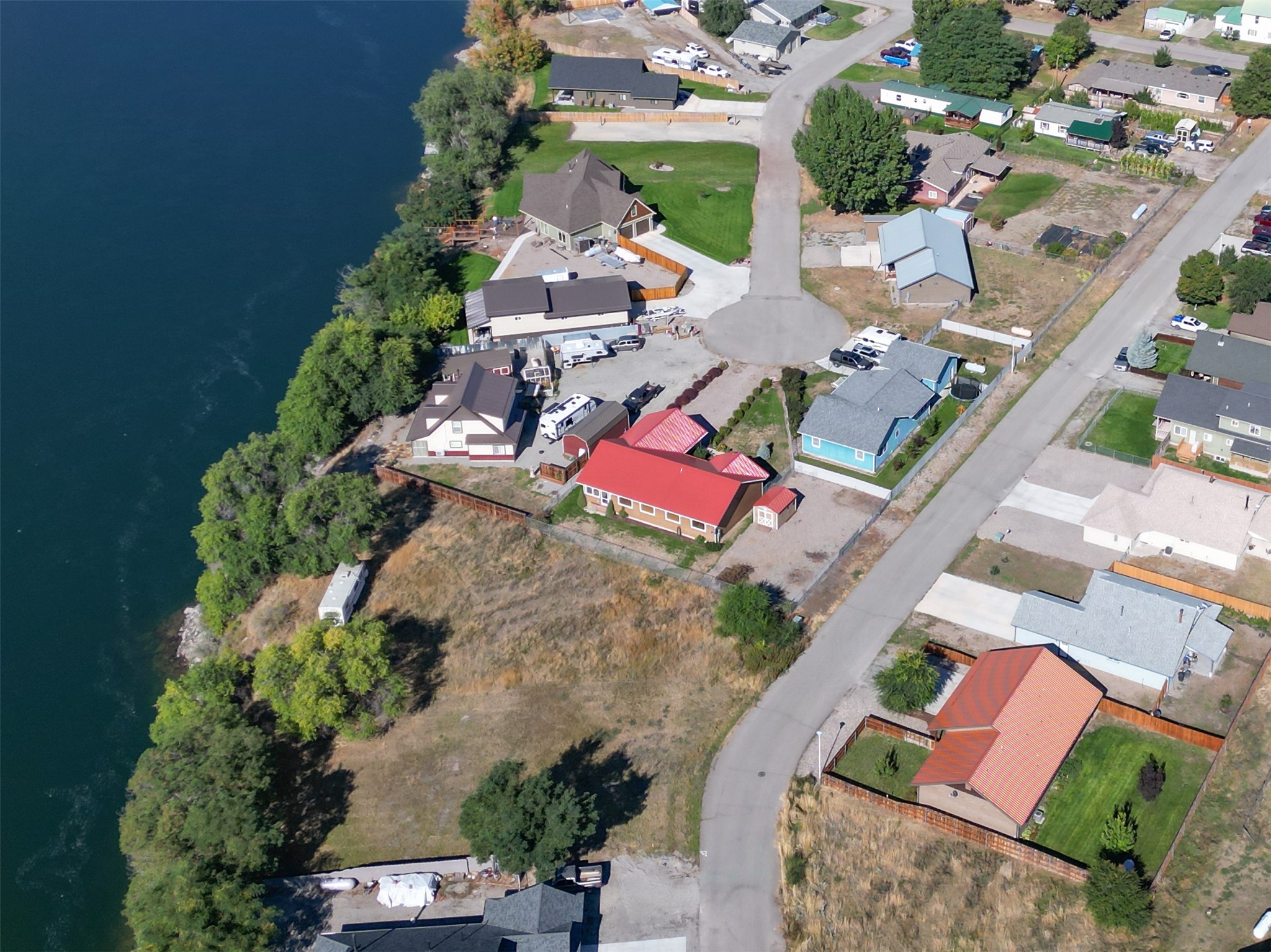 Aerial view of shoreline homes, a curving road, and a dry open lot beside the water.