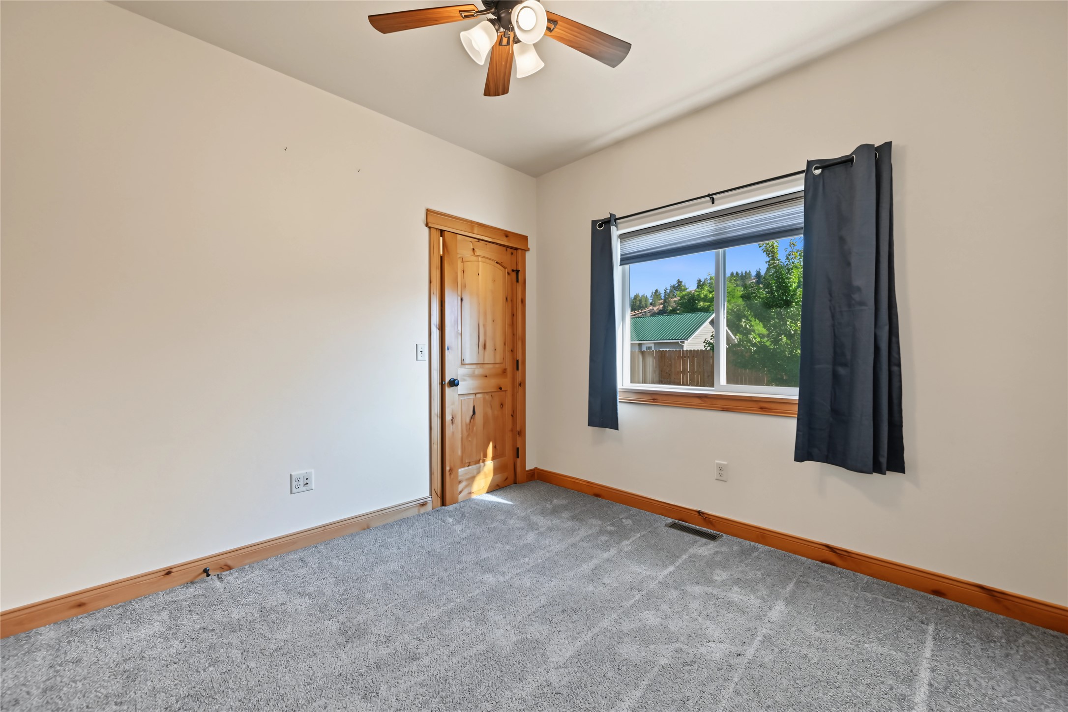 Empty carpeted room with wood trim, a wooden door, and a curtained window overlooking trees.