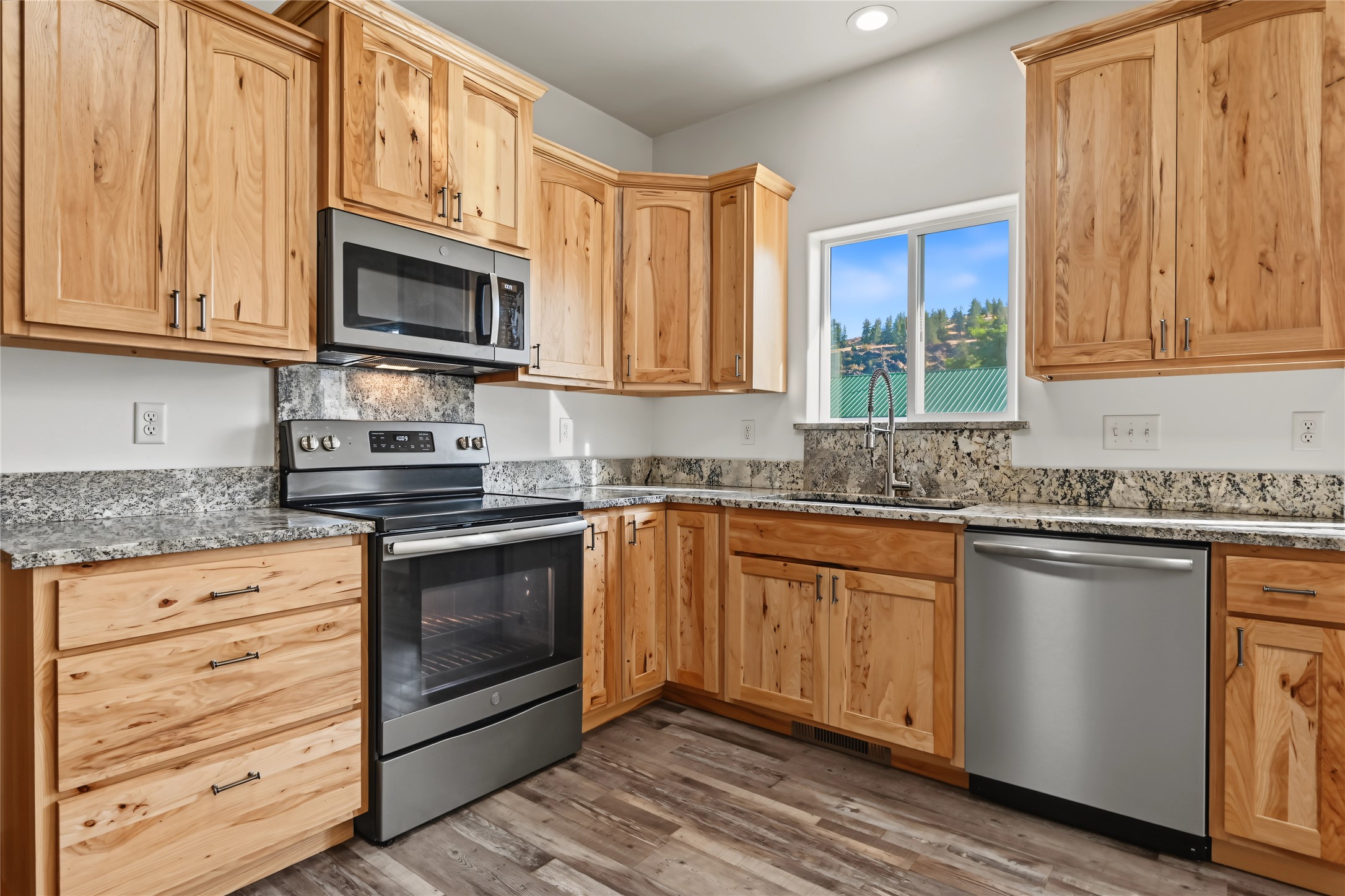 Kitchen with natural wood cabinets, speckled stone counters, stainless appliances, and a window overlooking a hillside.
