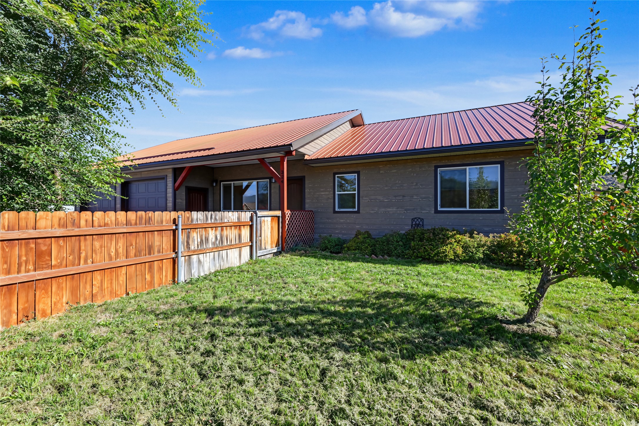 Single-story gray house with a red metal roof, attached garage, and fenced grassy yard.