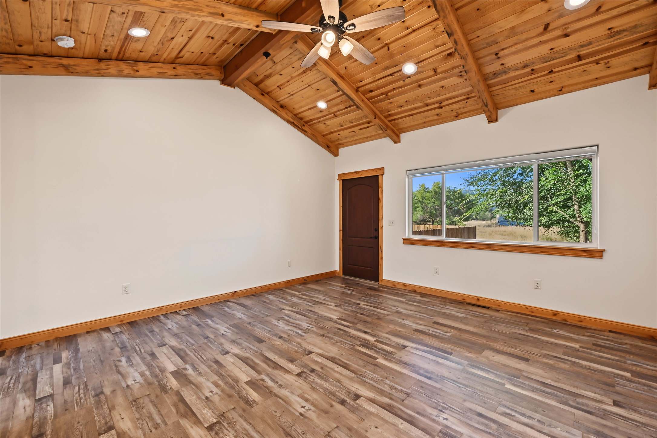 Empty room with a vaulted pine ceiling, exposed beams, and a wide window overlooking trees.