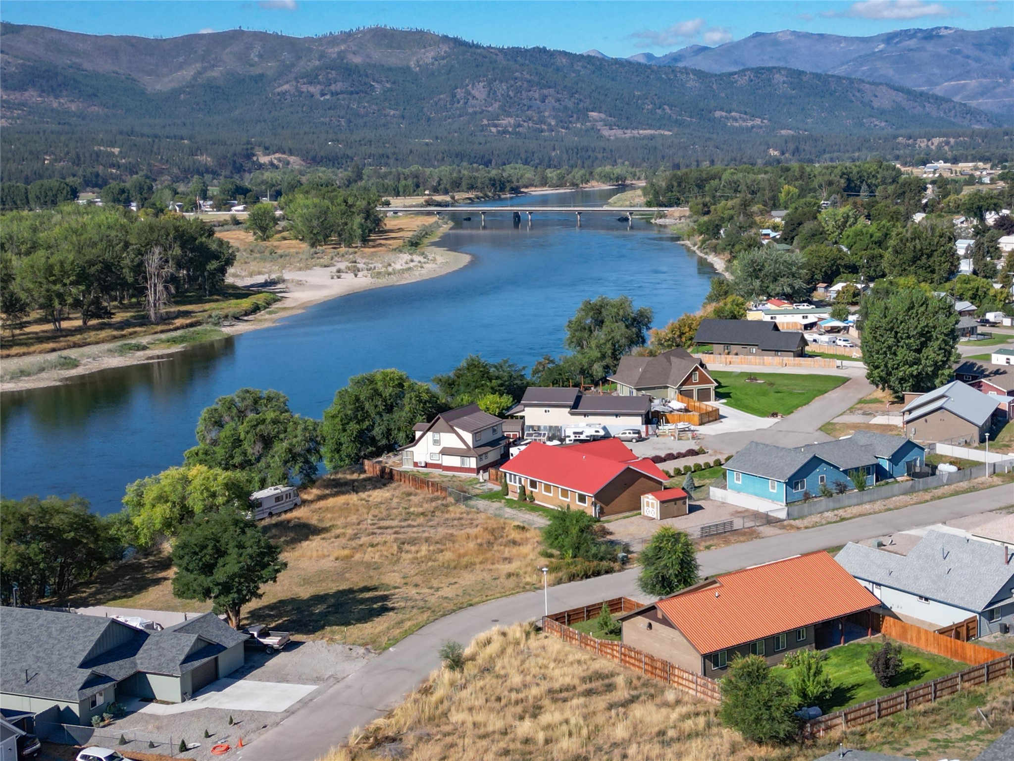 Aerial view of homes beside a river, with a bridge and forested mountains beyond.