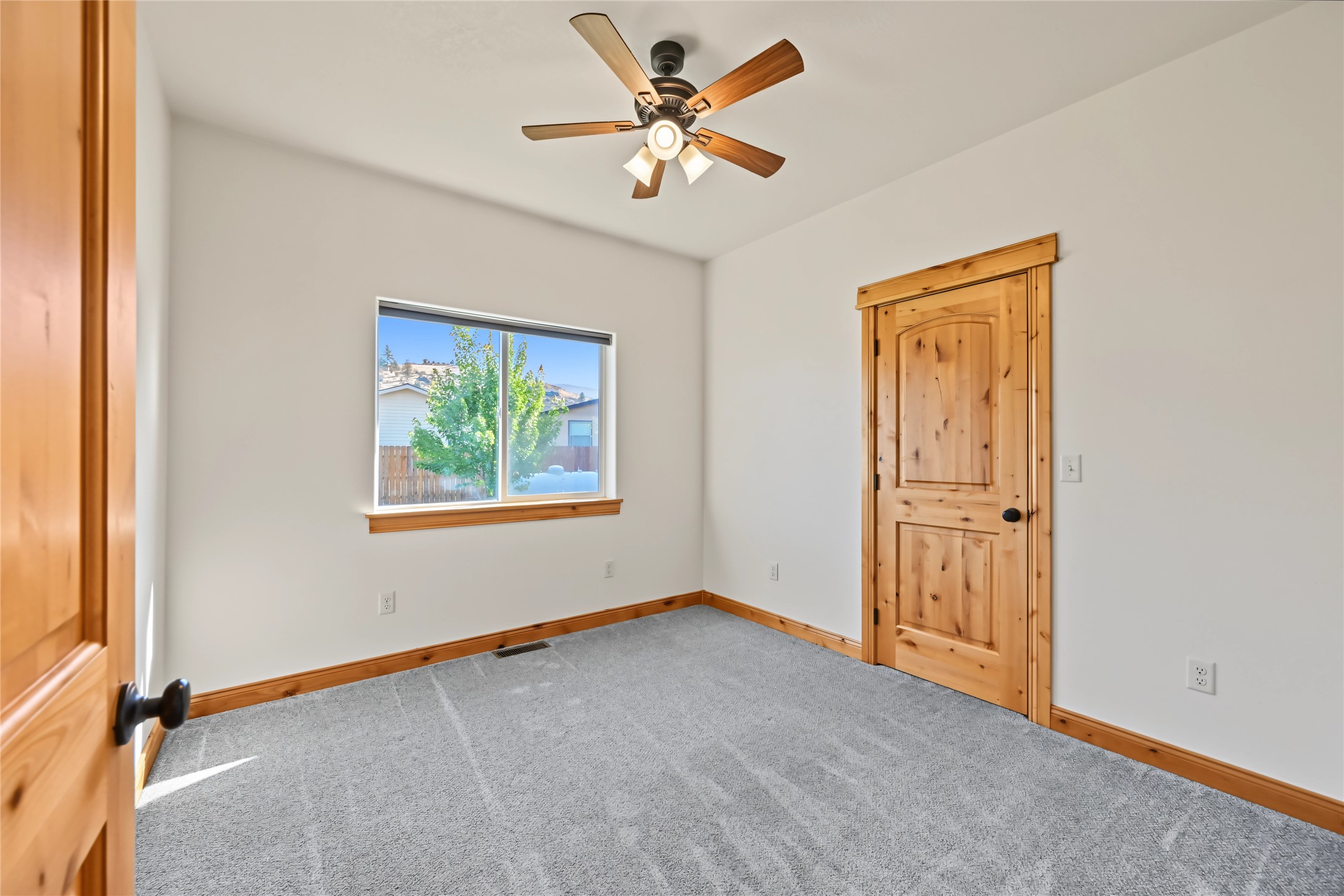 Empty bedroom with gray carpet, wood trim, a ceiling fan, and a window overlooking a tree.