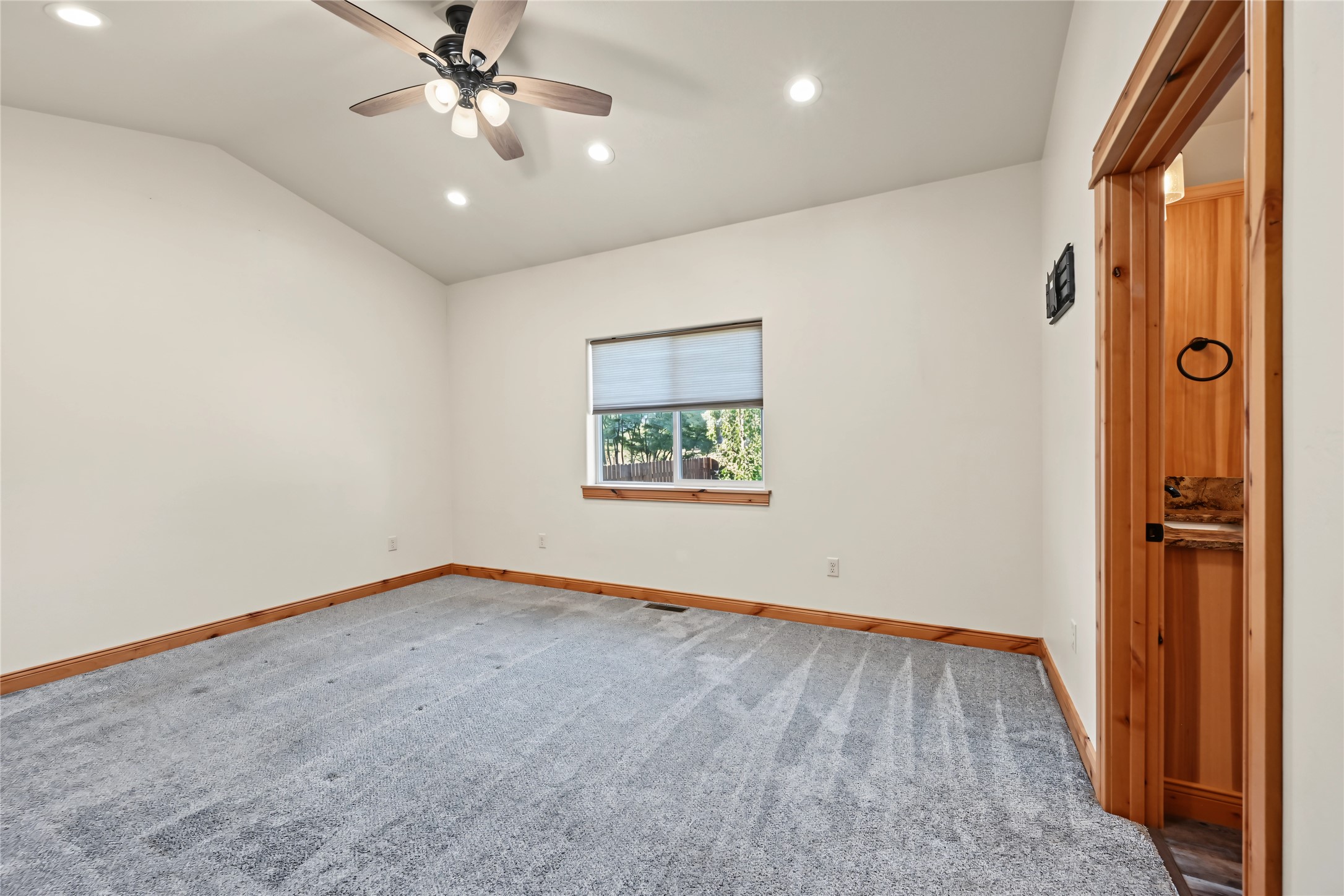 Empty bedroom with gray carpet, wood trim, ceiling fan, and a window with a partially lowered blind.