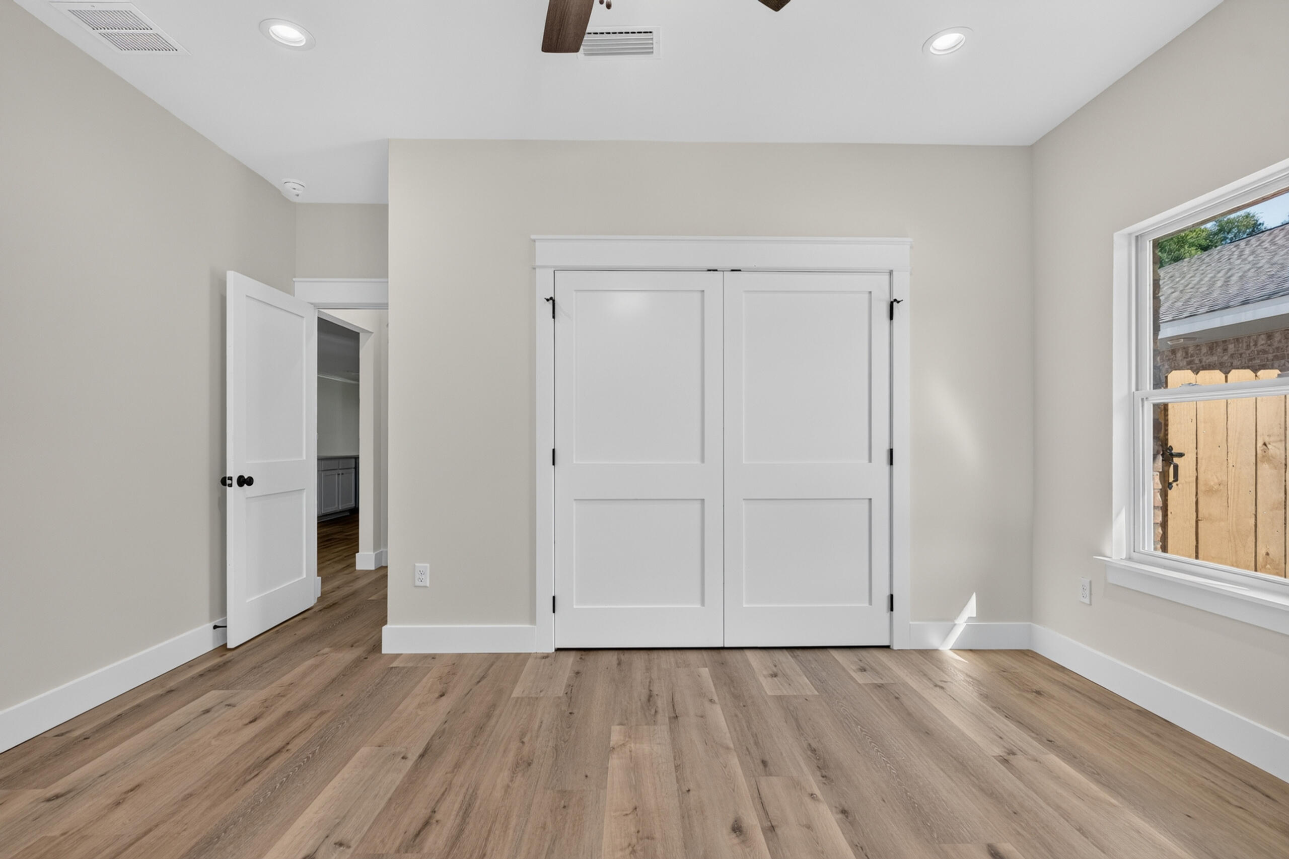 Empty bedroom with wood-look floors, double closet doors, and a window facing a wooden fence.