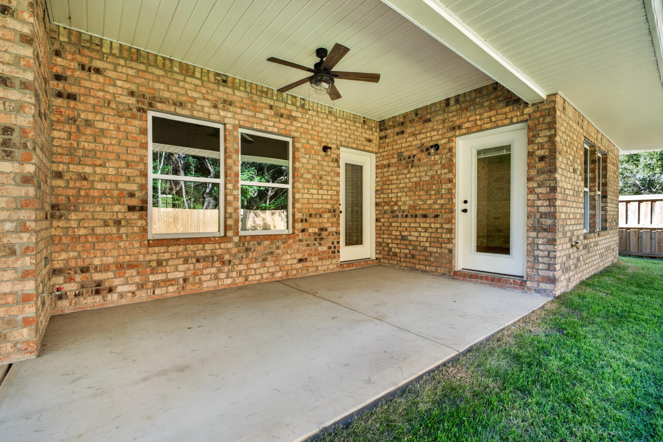 Covered brick patio with a ceiling fan, two windows, and white doors beside a grassy yard.