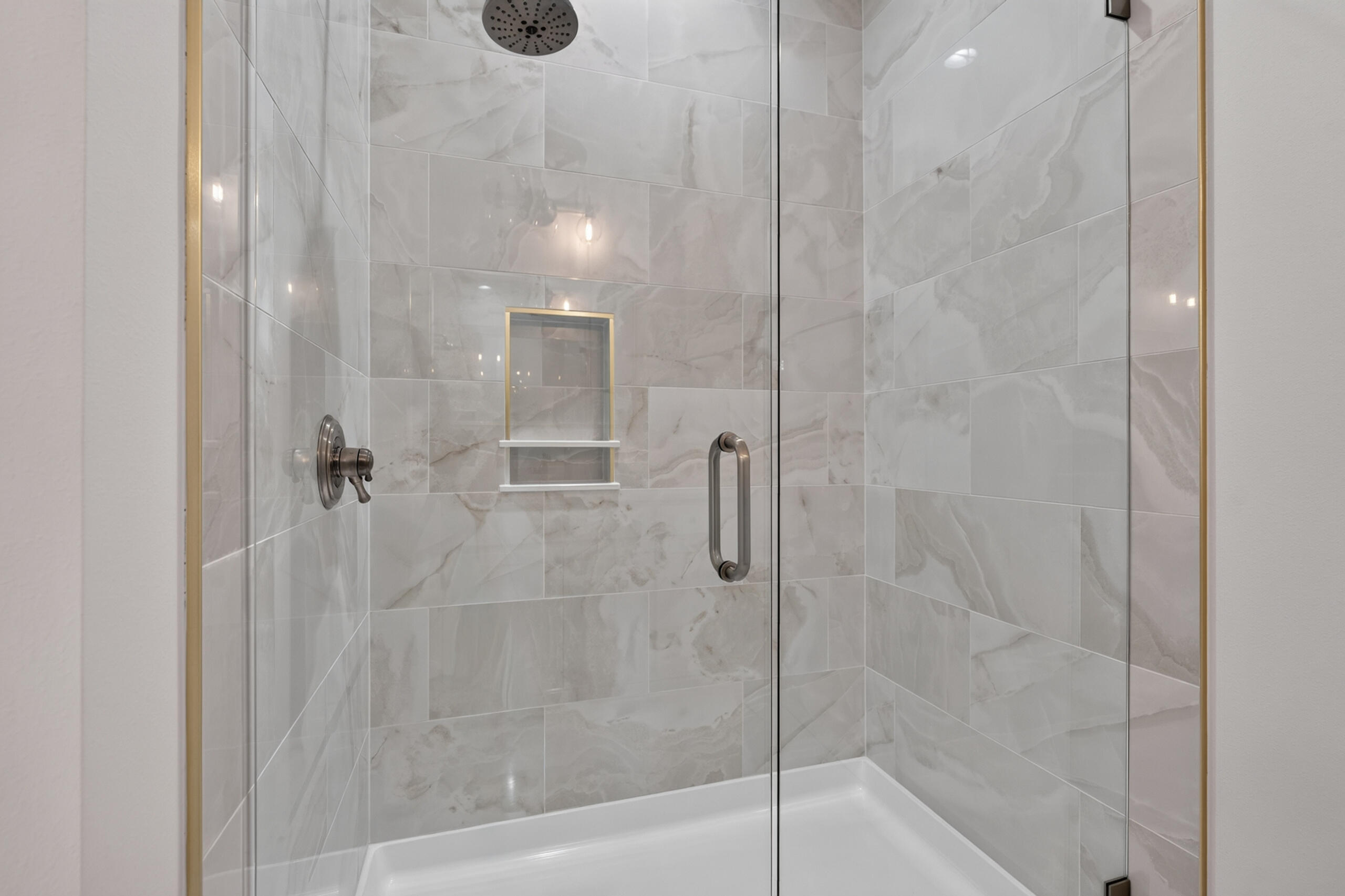 Walk-in shower with marble-look tile, glass doors, rainfall showerhead, and recessed shelf.