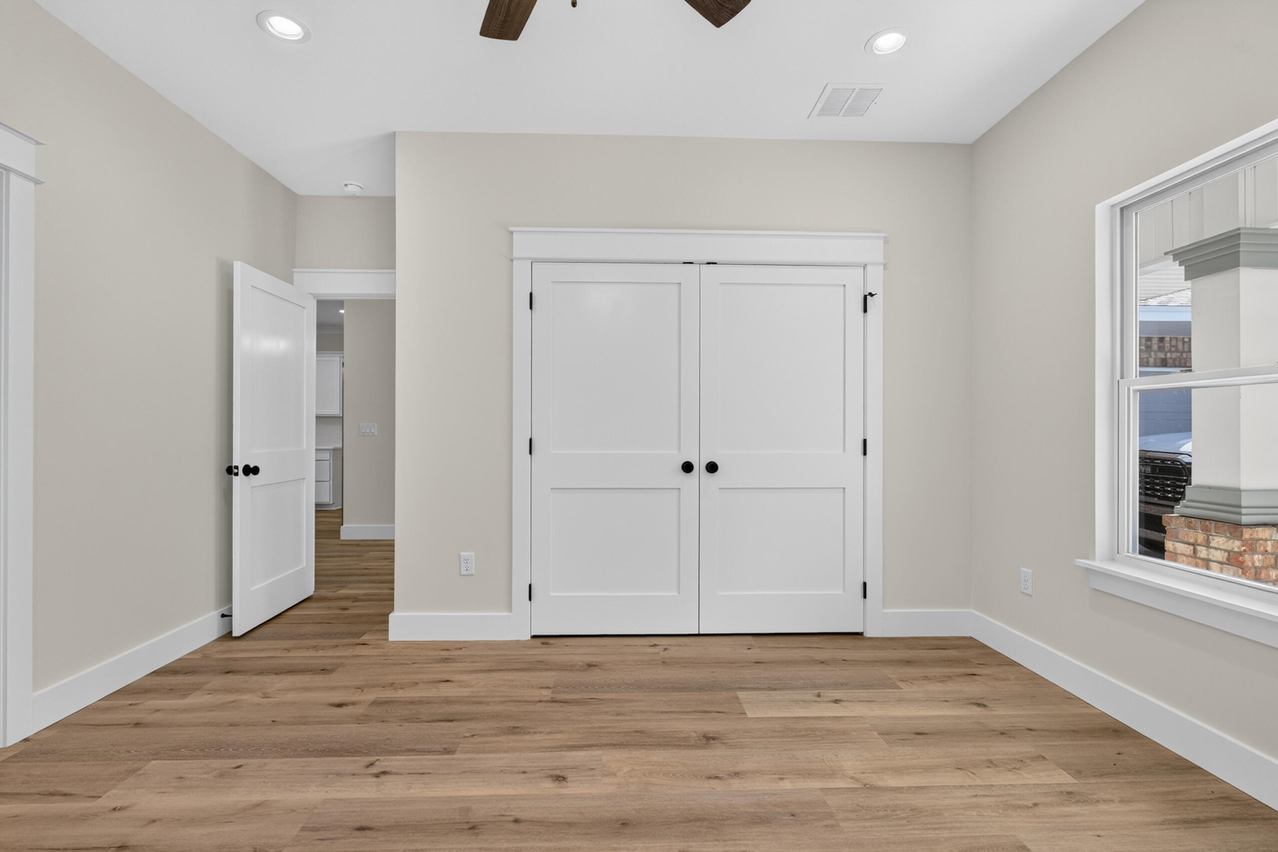 Empty bedroom with wood flooring, white double closet doors, and a large window.