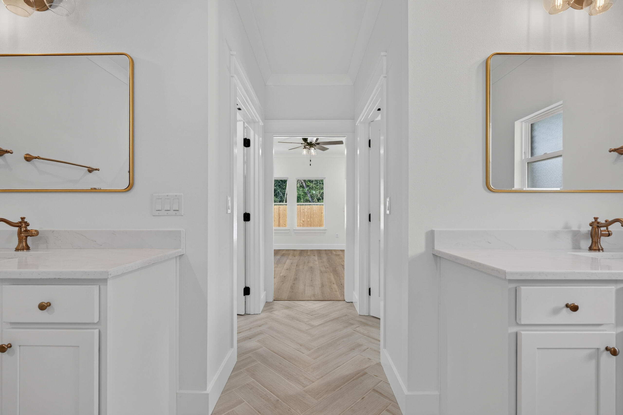 Bathroom with matching white vanities and brass fixtures, looking through a tiled hall toward a bedroom.